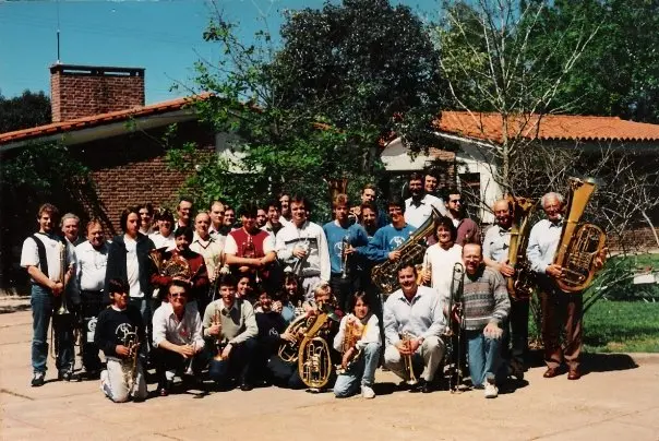 Unas treinta y cinco personas con instrumentos de bronce posando al aire libre frente a una casa de ladrillo con techo de tejas, con dos tubas grandes a la derecha