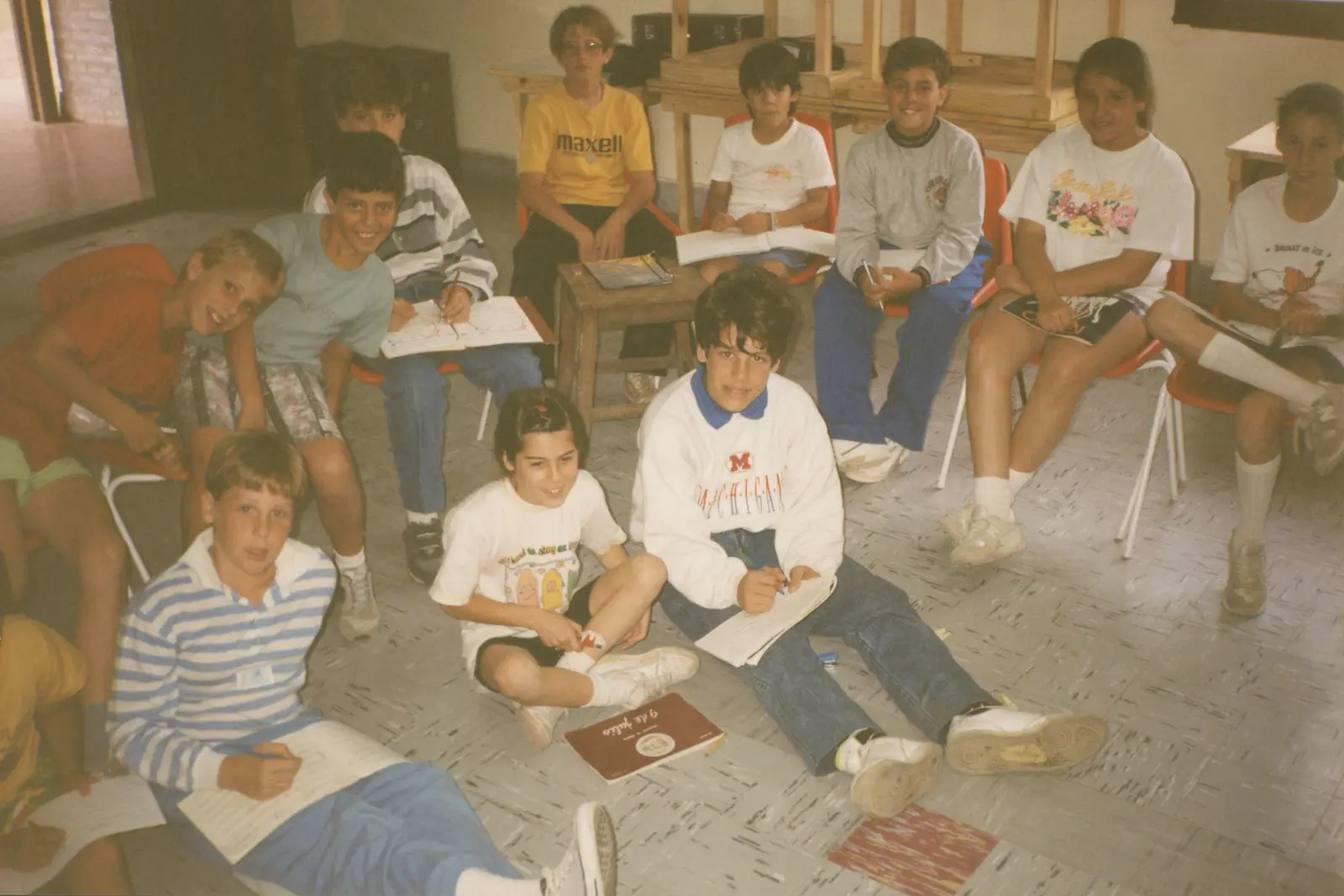 Trece chicos y chicas en una sala, unos en sillas de plástico rojas y otros sentados en el piso, escribiendo con birome en cuadernos de pentagramas