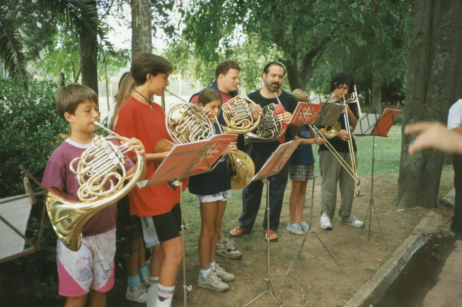 Fila de músicos tocando al aire libre bajo los árboles: tres chicos y dos adultos con cornos franceses y un adolescente con trombón, frente a atriles con carpetas rojas