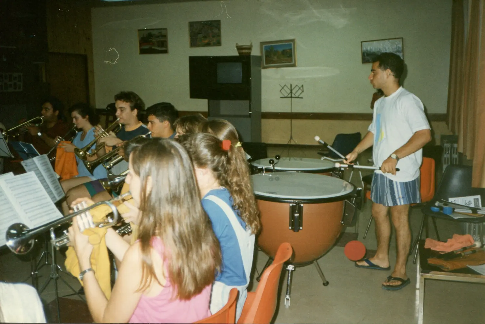 Un joven de pie junto a dos timbales, con las baquetas en alto, frente a un grupo de chicos sentados tocando trompetas