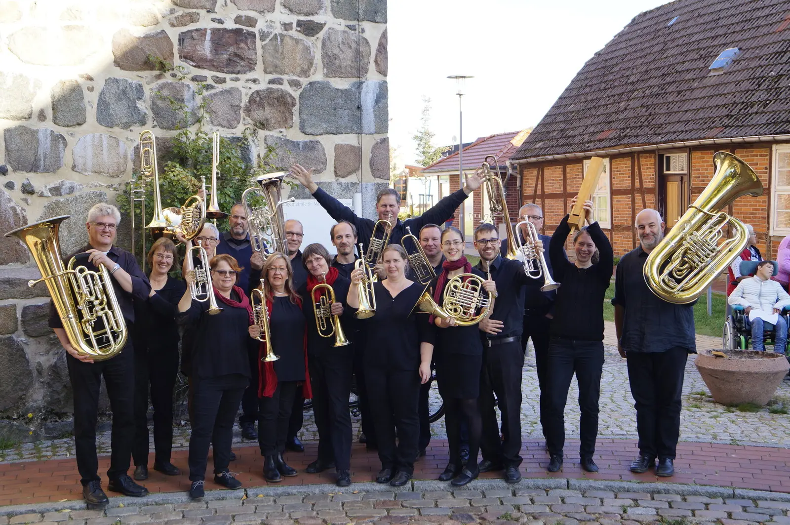 Los dieciocho músicos del Bläserkreis M-V de negro, con los instrumentos en alto, delante de una iglesia de piedra y una casa de entramado