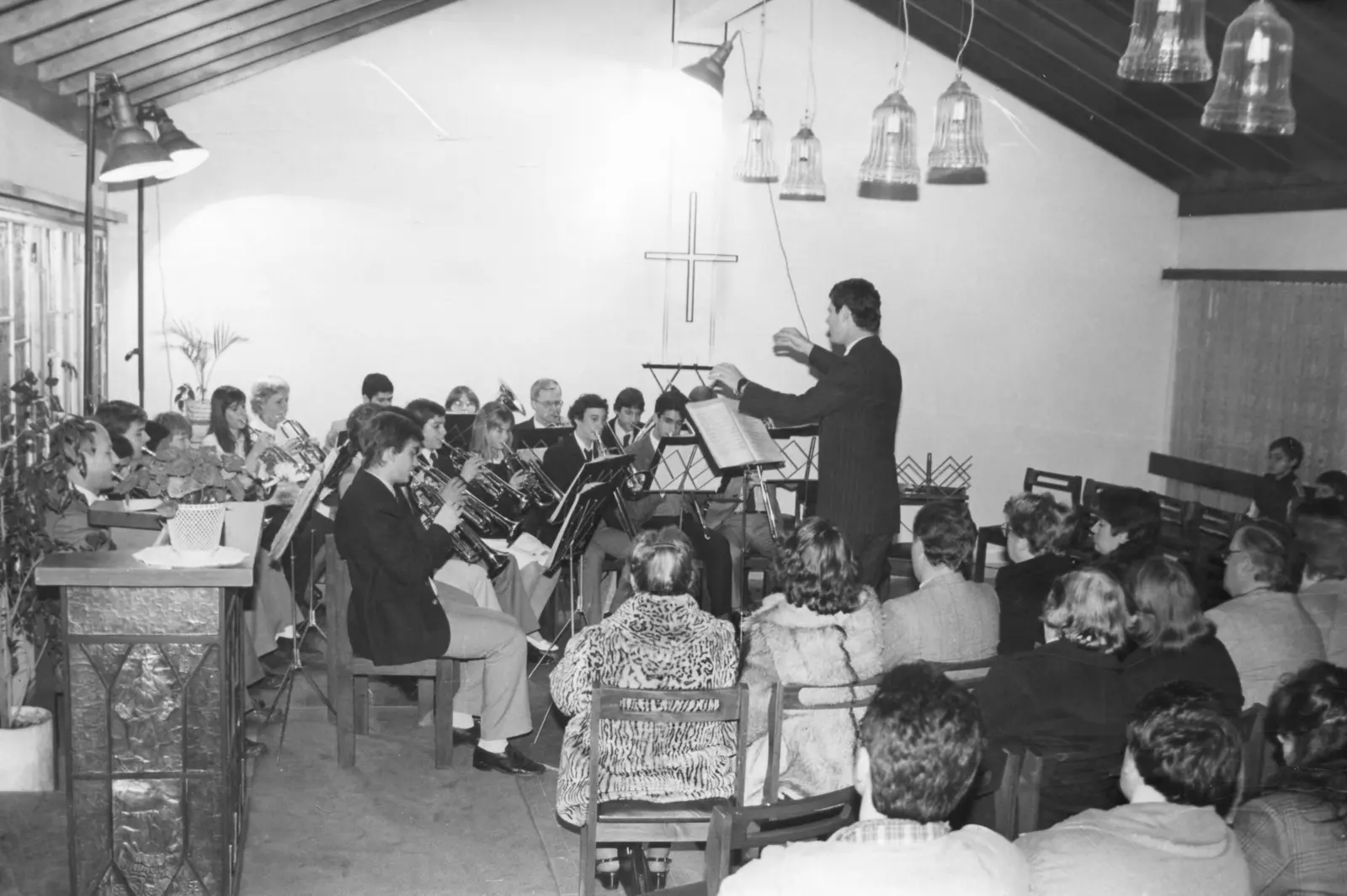 Fotografía en blanco y negro del interior de una capilla pequeña de techo a dos aguas con vigas de madera y lámparas colgantes de vidrio: unos quince músicos sentados a la izquierda tocando y el público sentado a la derecha, varias mujeres con tapados de piel
