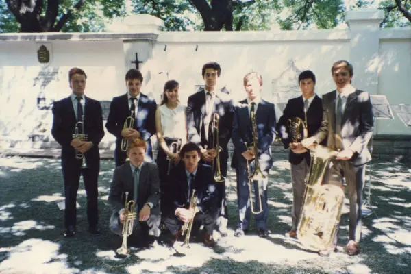 Nueve músicos de traje con sus instrumentos posando frente al muro blanco del Cementerio Alemán, principios de los noventa