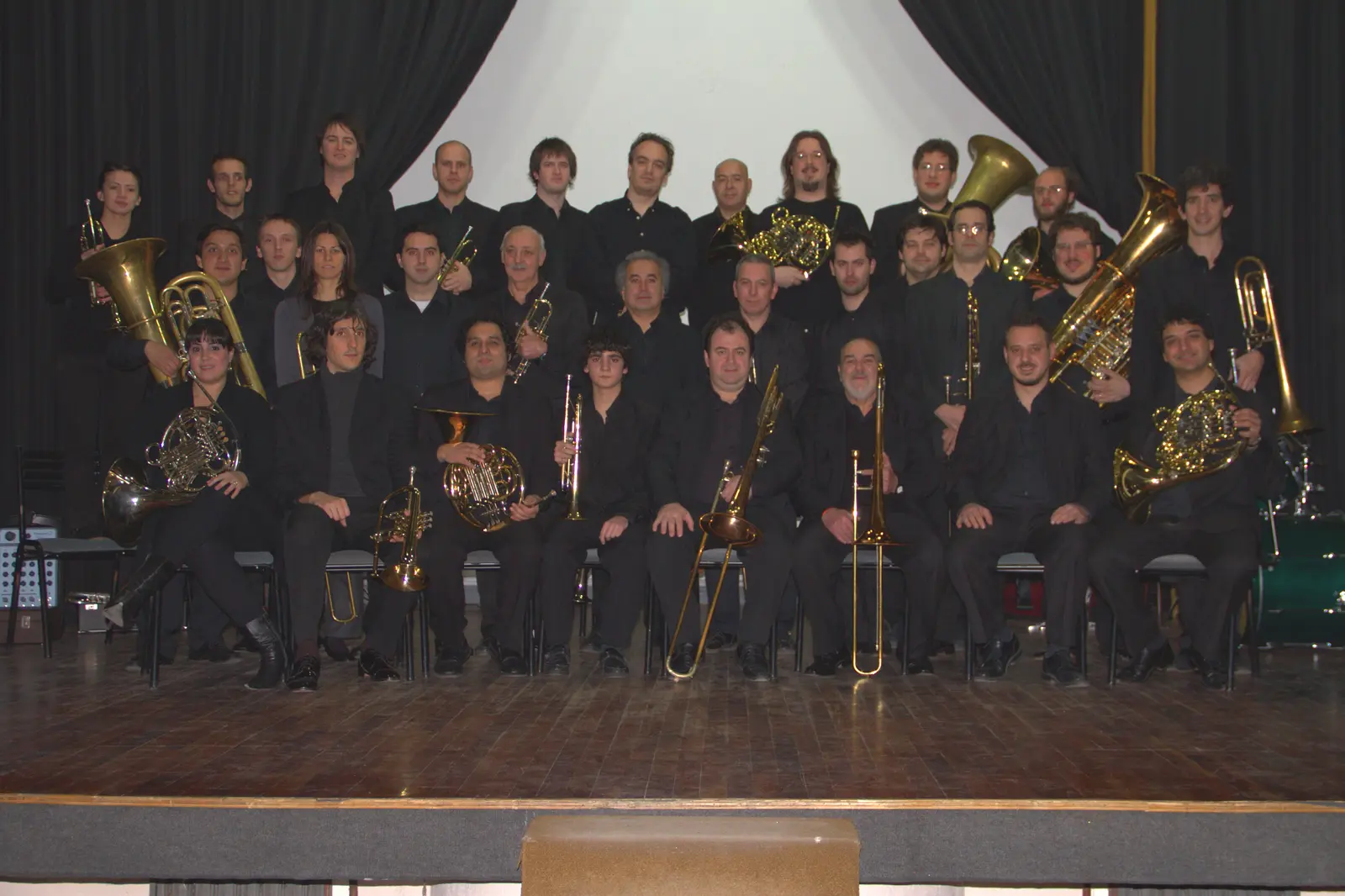Retrato formal de unos treinta y cinco músicos de negro en tres filas sobre un escenario, con tubas, cornos, trombones, bombardinos y trompetas
