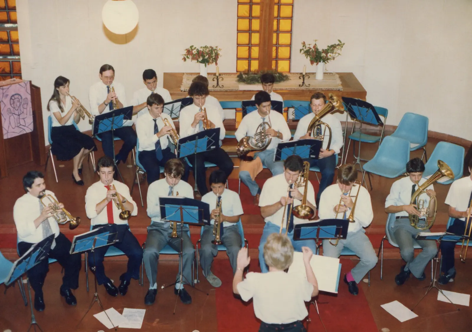 Vista elevada del coro tocando dentro de una iglesia moderna: dos semicírculos de sillas celestes con unos diecisiete músicos de camisa blanca y corbata, atriles azules, y al fondo el altar de madera con una cruz sobre un vitral de bloques anaranjados