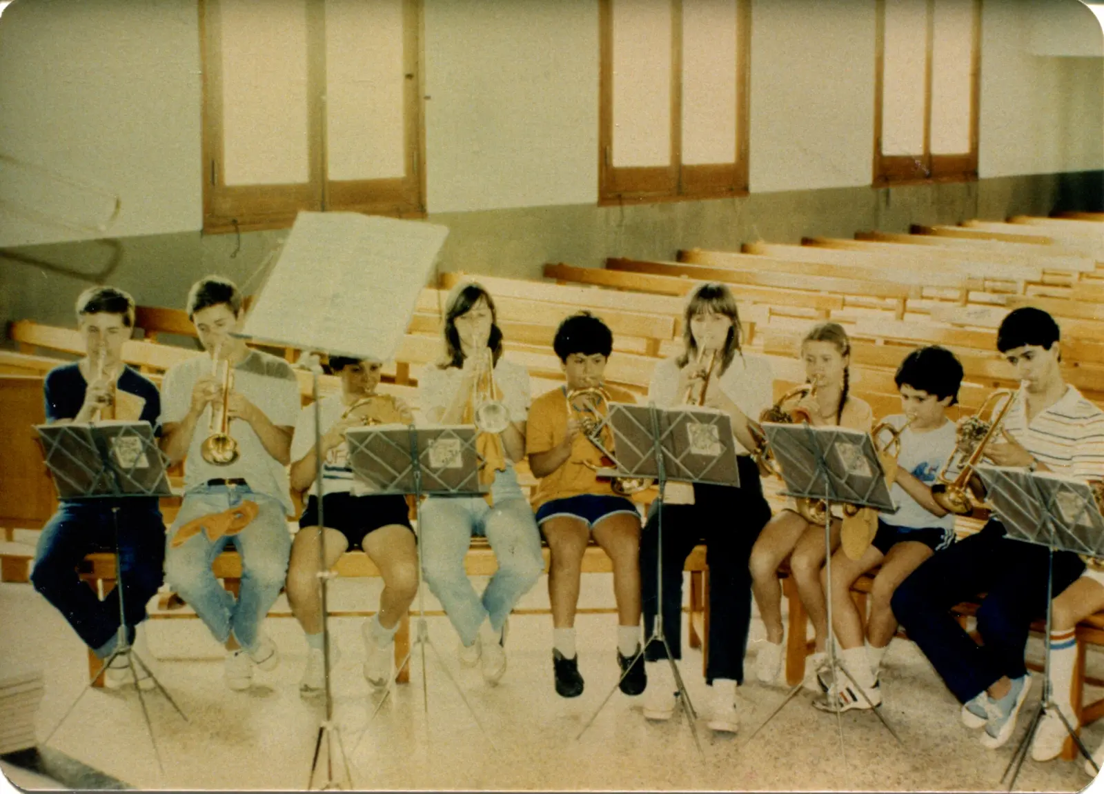 Diez adolescentes sentados en fila sobre bancos de madera clara, ensayando con trompetas, cornos y un trombón dentro de un templo vacío, todos en ropa de verano: shorts, jeans y zapatillas