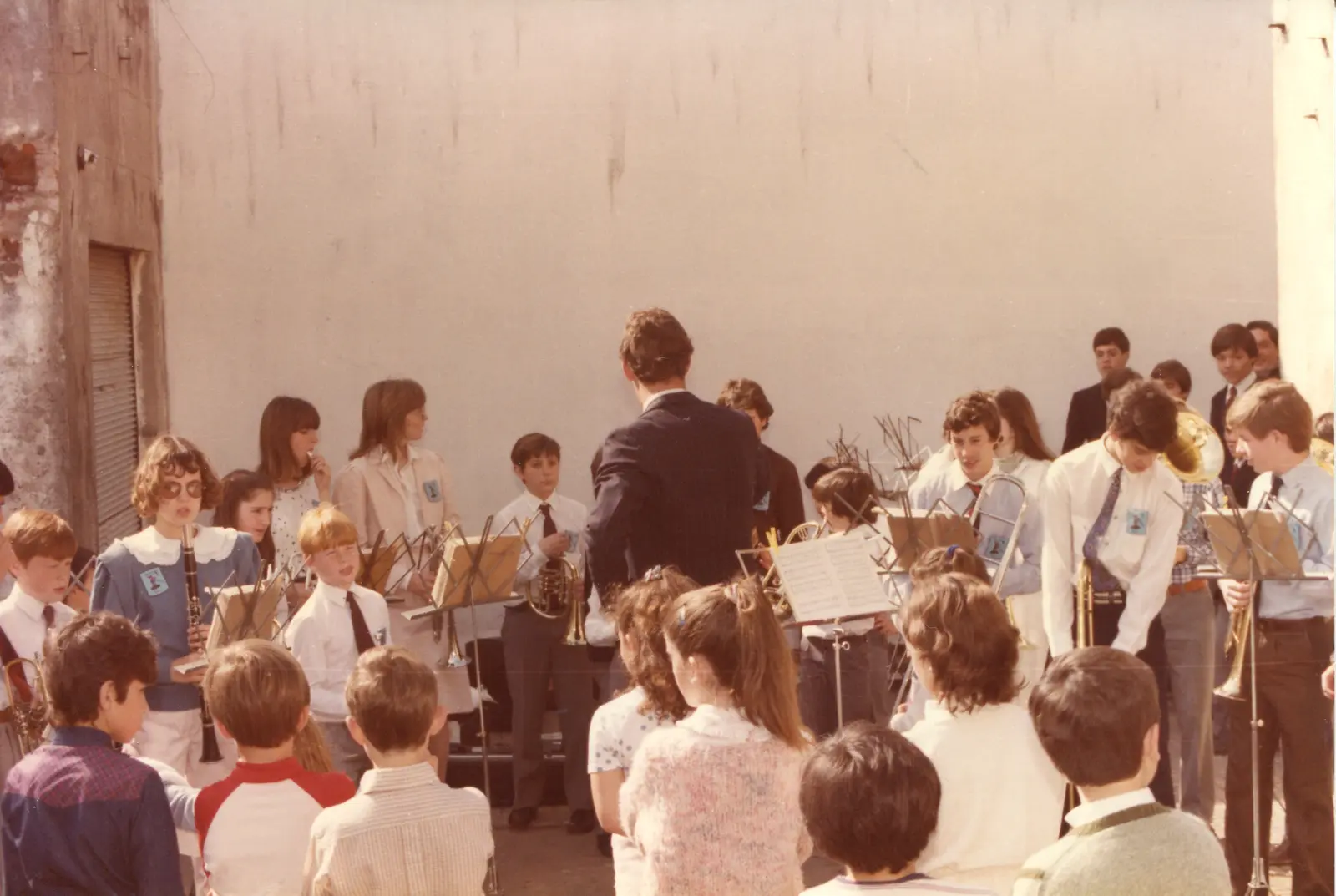 Concierto en un patio contra una pared blanca alta con persiana metálica: unos veinte chicos y adolescentes tocando frente a atriles, el director de espaldas con traje azul, y varios con un escudo claro cosido en el pecho