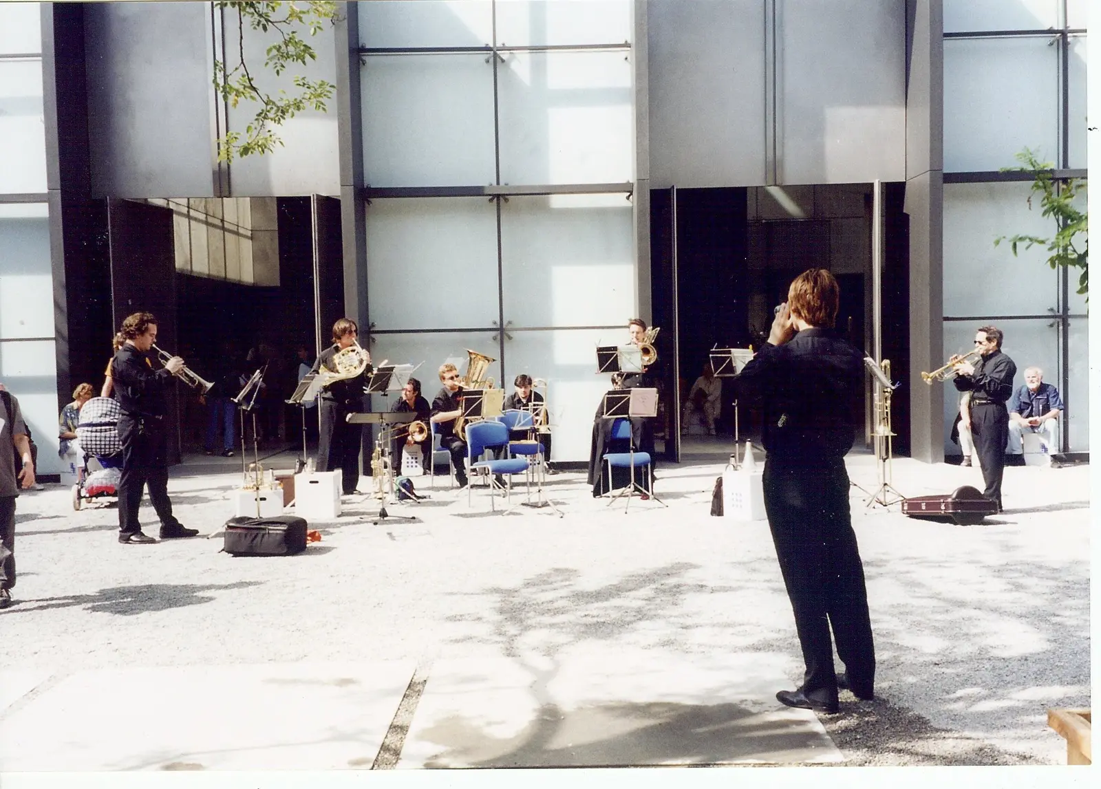 El coro de negro tocando al aire libre en el patio de gravilla de un edificio moderno de vidrio y acero, con el director de espaldas y público sentado a la sombra