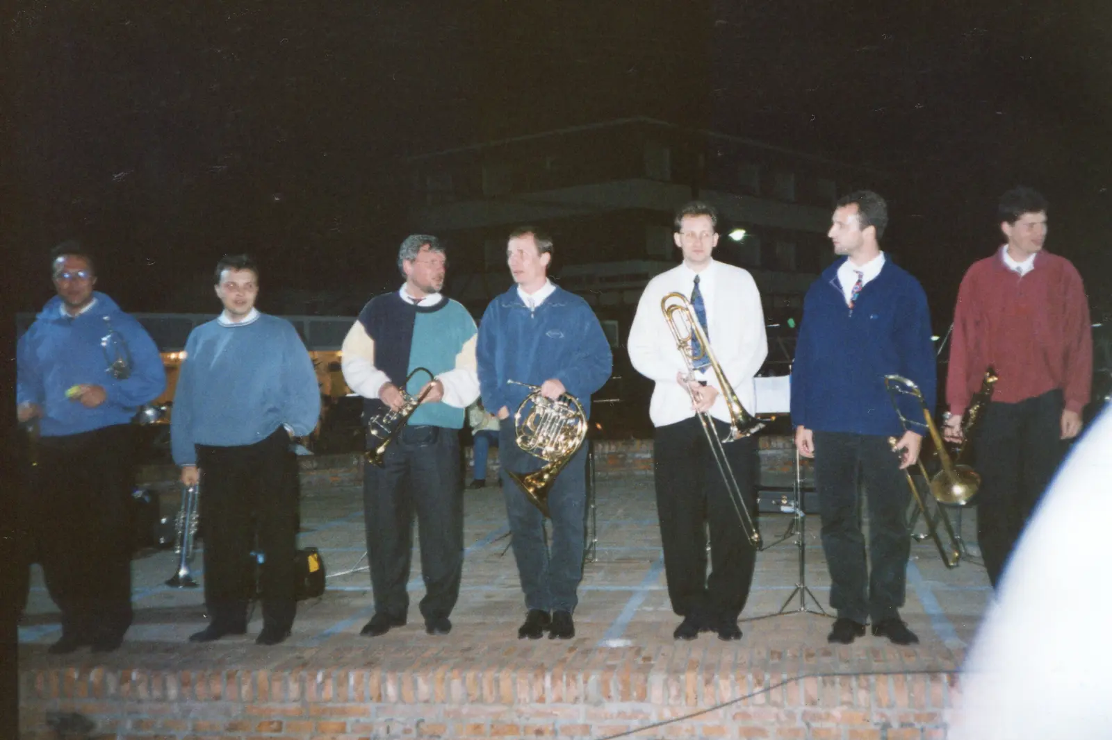 Ocho músicos de pie sobre una tarima de ladrillo, de noche, al aire libre, con un edificio iluminado detrás