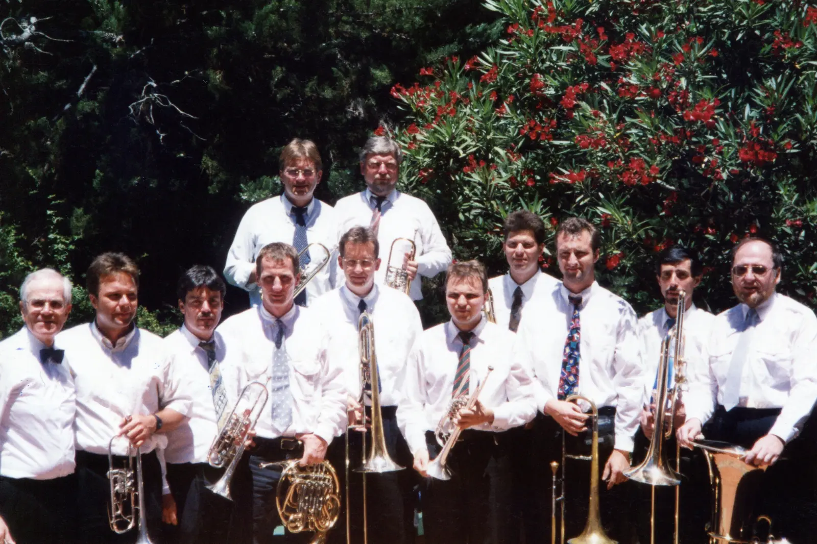 Trece músicos alemanes de camisa blanca posando con sus instrumentos frente a un arbusto de flores rojas