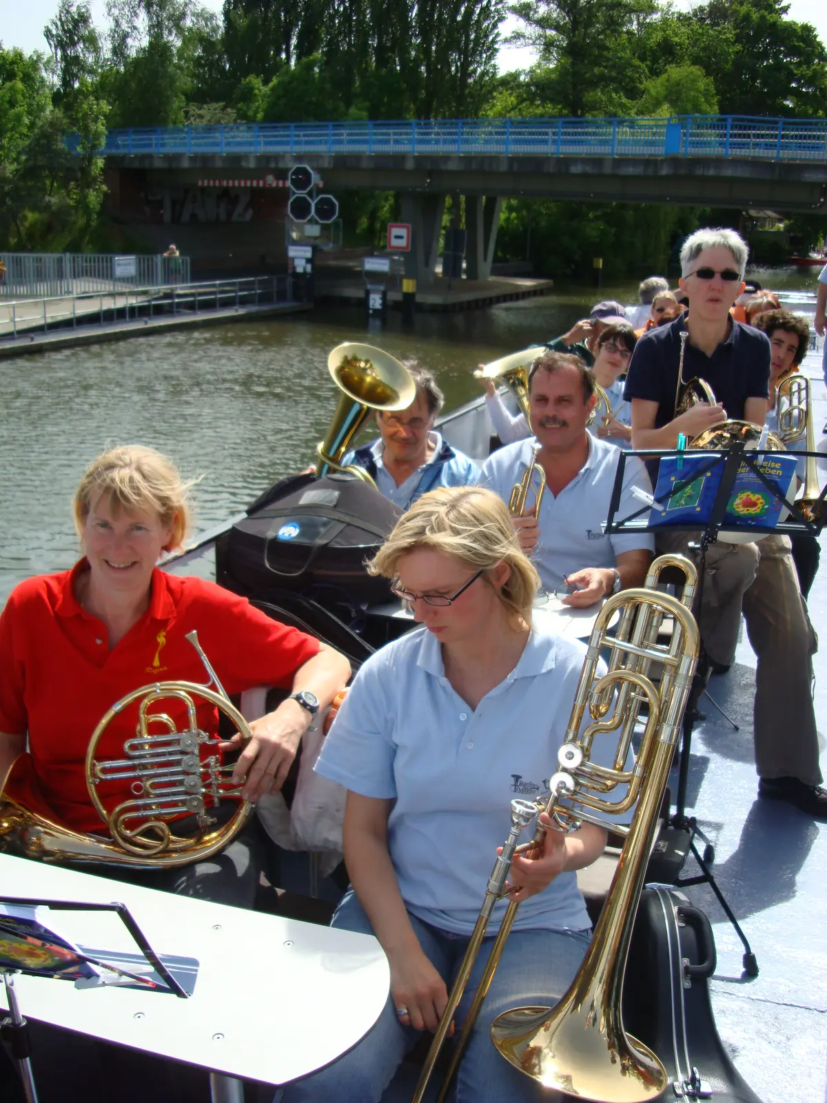 Músicos sentados con sus instrumentos en la cubierta abierta de un barco que pasa bajo un puente metálico azul, en un canal bordeado de árboles