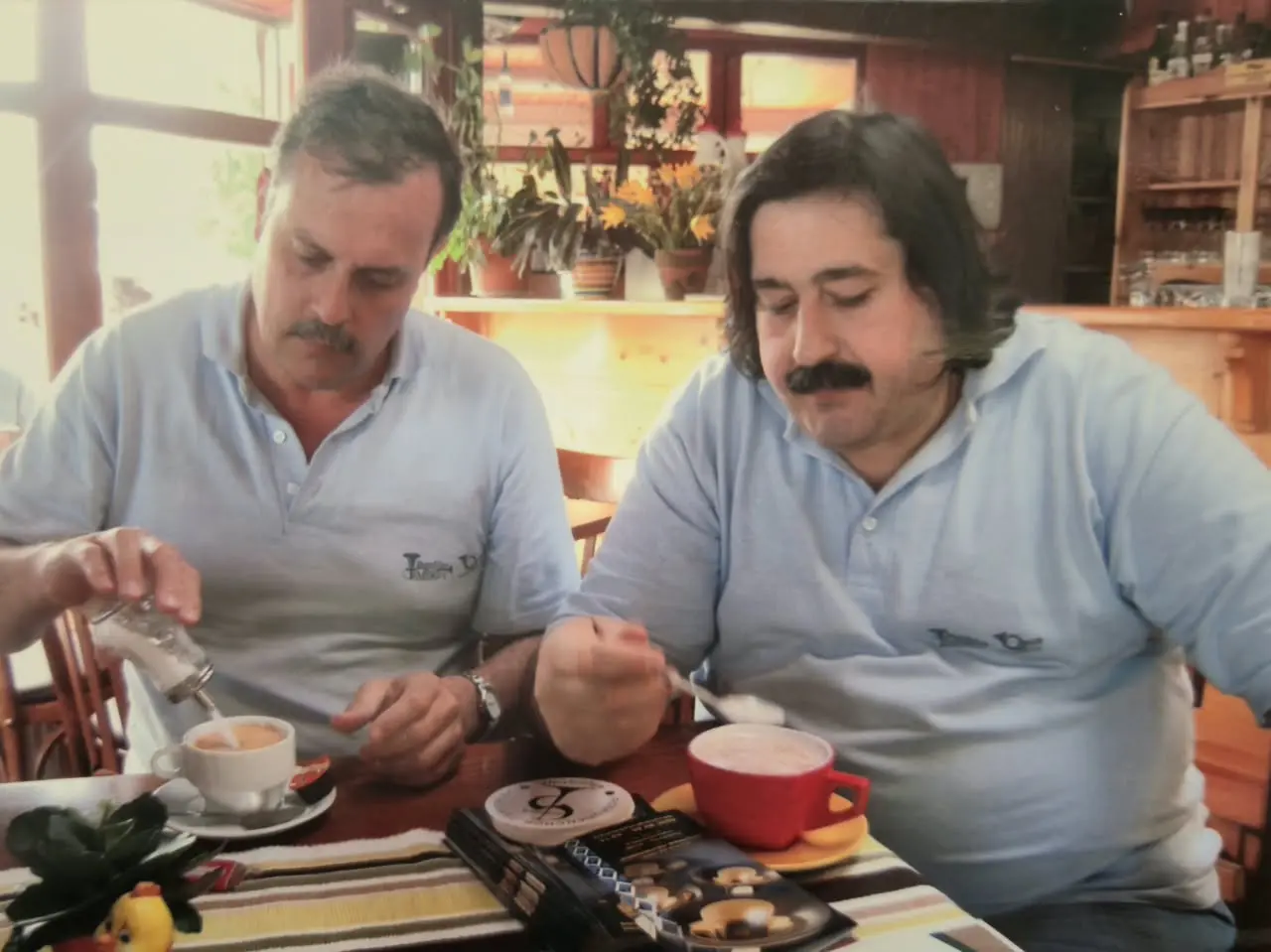 Dos integrantes de la gira con la chomba celeste desayunando en un salón de madera, uno echando azúcar en un pocillo y el otro revolviendo una taza roja