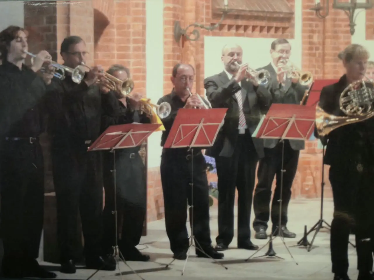 Siete músicos tocando de pie ante atriles rojos dentro de una iglesia de ladrillo visto, cuatro de negro y tres de traje, con una trompa al borde derecho