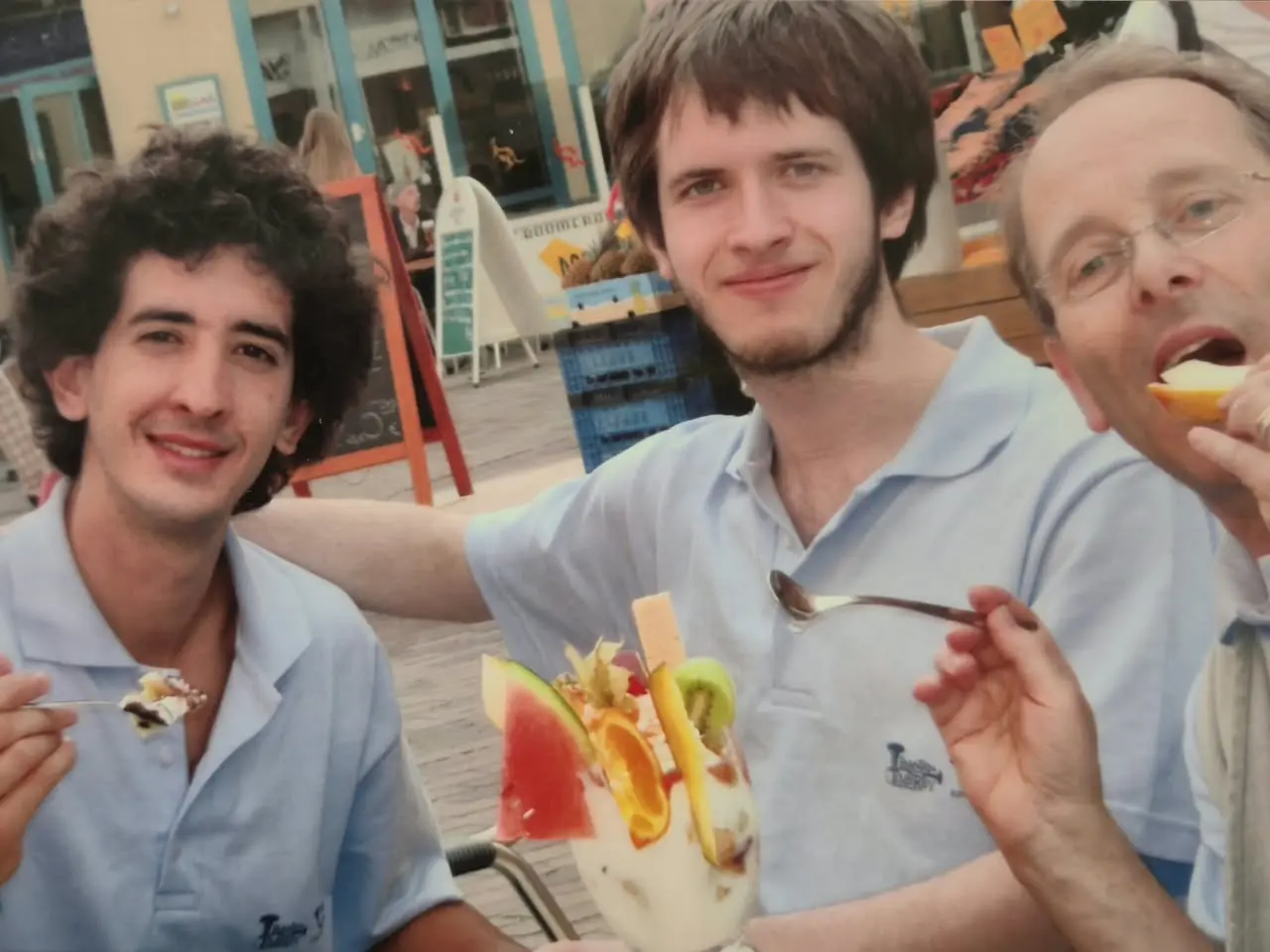 Tres integrantes de la gira con la chomba celeste del coro alrededor de una copa alta de helado con frutas, en la mesa de una heladería peatonal