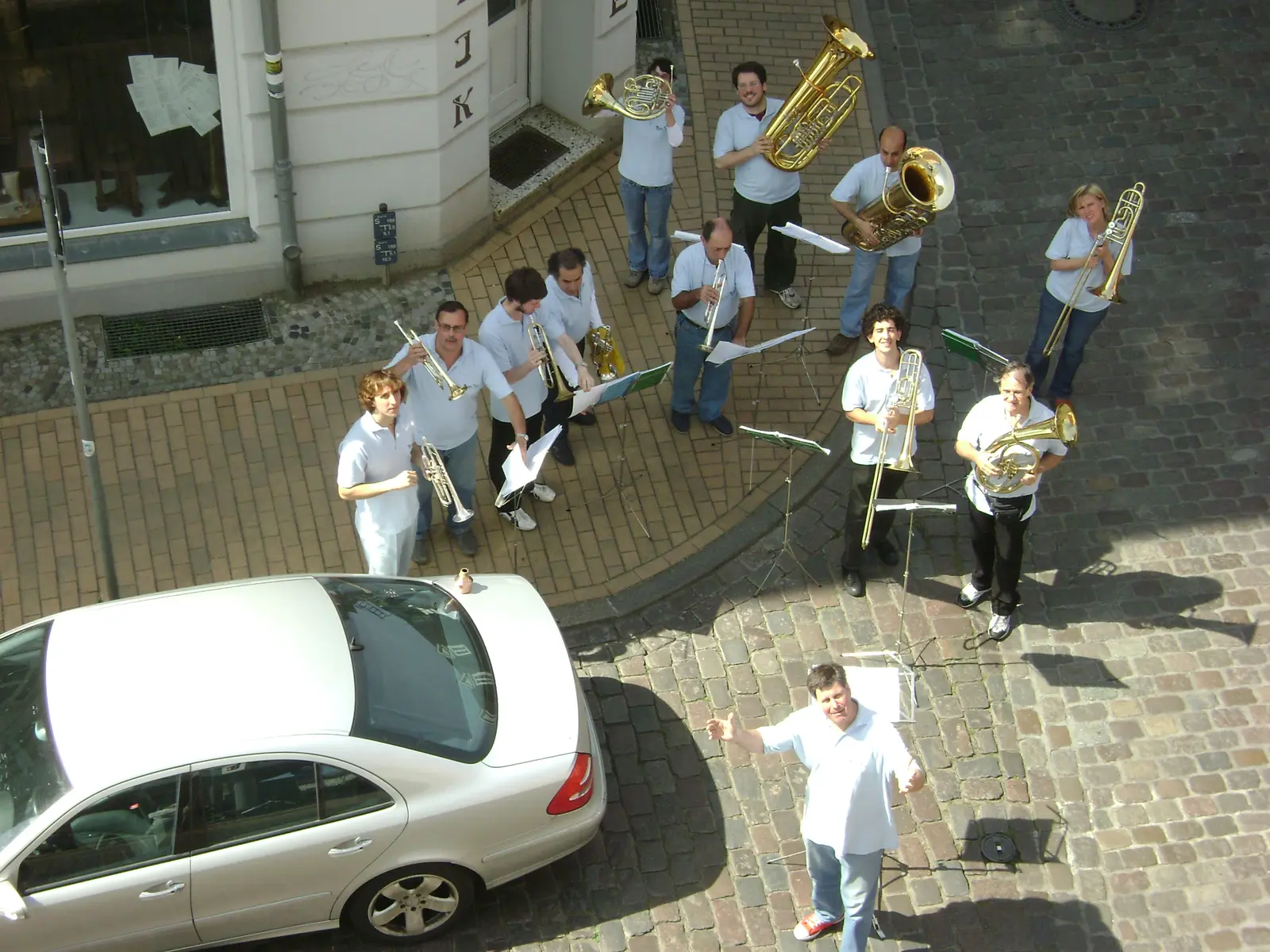 Vista desde una ventana alta: doce músicos con chombas celestes tocando en una esquina empedrada, con atriles y un Mercedes estacionado; el director, abajo, abre los brazos mirando a la cámara
