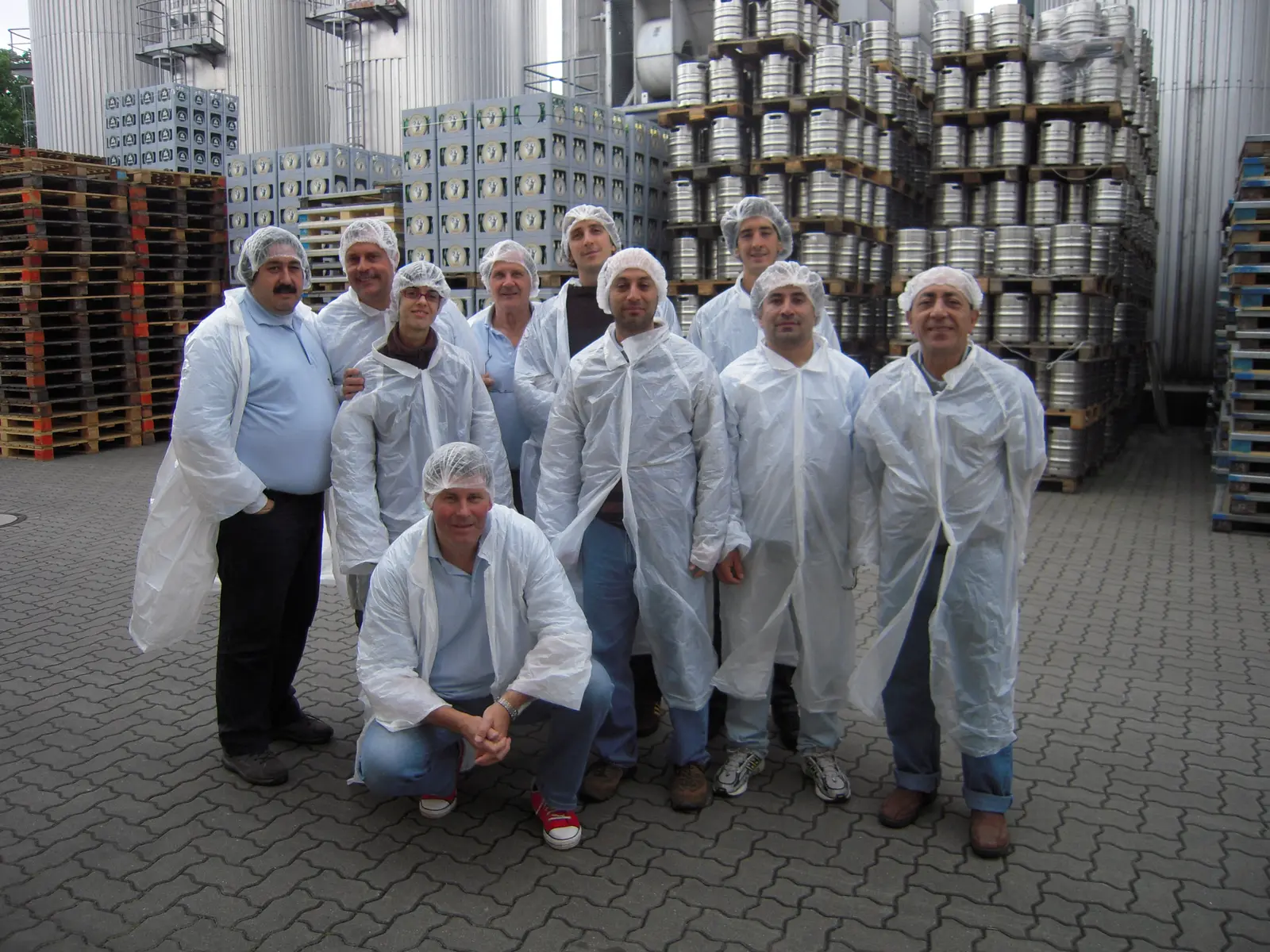 Once integrantes posando con delantales blancos descartables y cofias en el patio de una cervecería, entre silos de acero, pilas de barriles metálicos y cajones de botellas