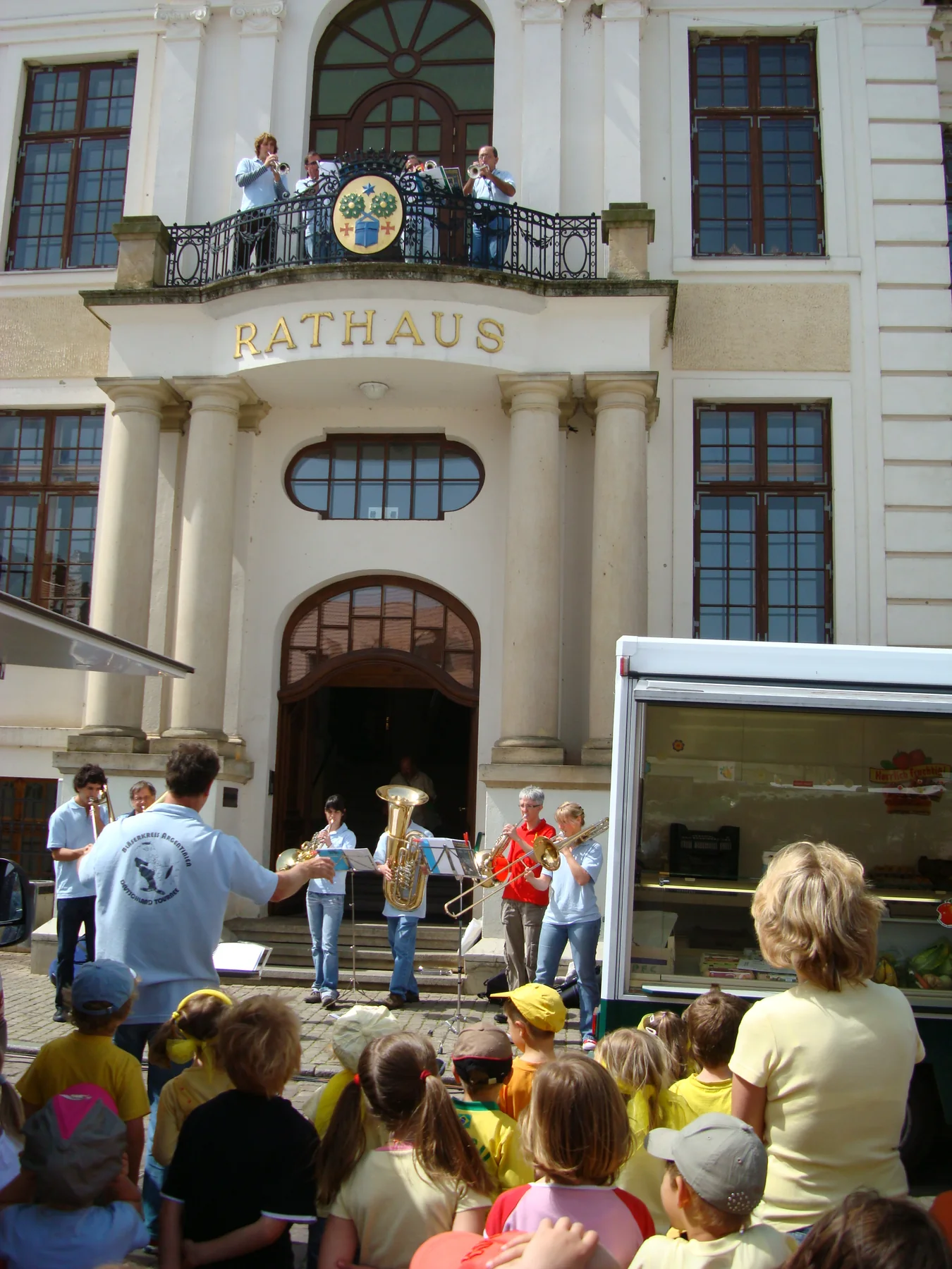 El conjunto tocando ante chicos en la plaza, con otros músicos en el balcón del Rathaus de Teterow