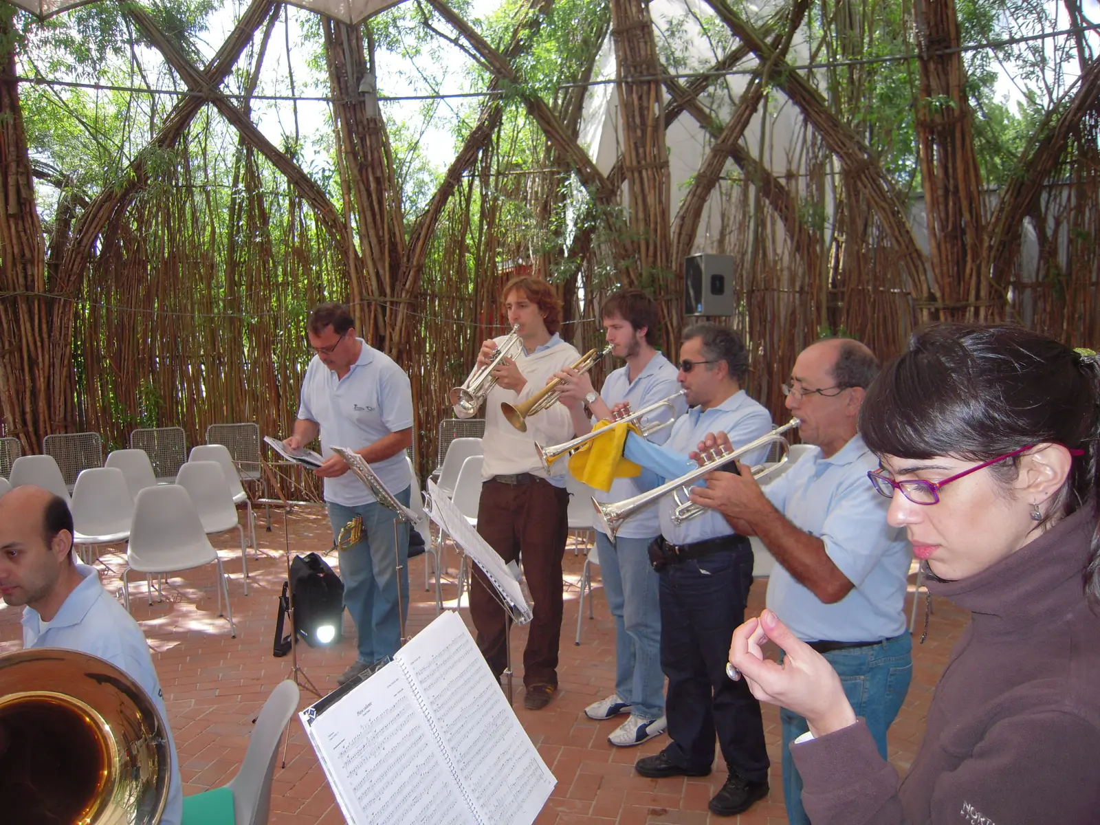 Músicos con chombas celestes tocando dentro de una cúpula formada por varas de sauce vivas entrelazadas en arcos, con sillas blancas de plástico y el cielo filtrándose entre las ramas