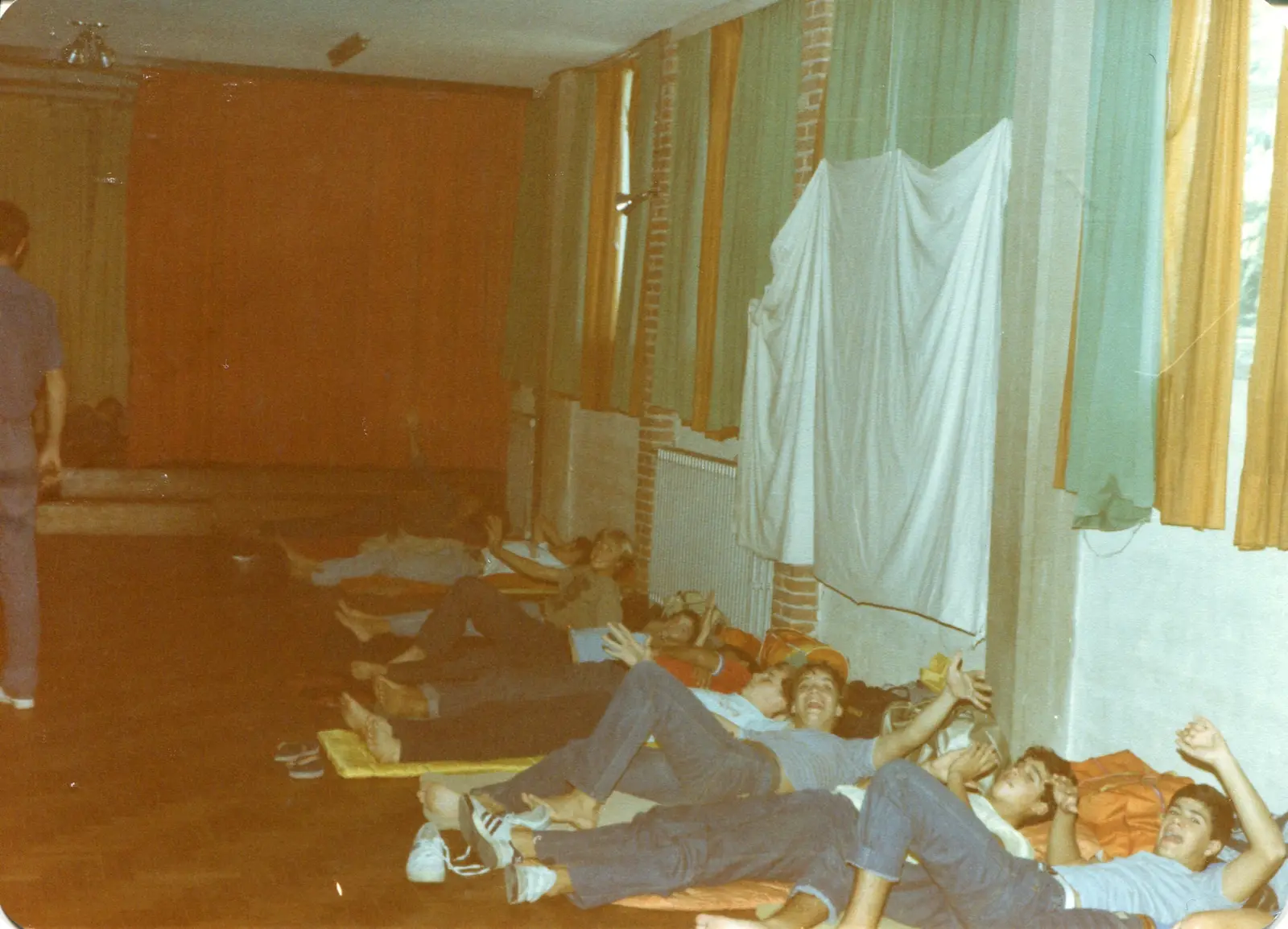 Una hilera de unos diez chicos y chicas acostados en el piso de madera de un salón grande, sobre colchonetas finas y bolsos, junto a una pared con cortinas verdes y amarillas