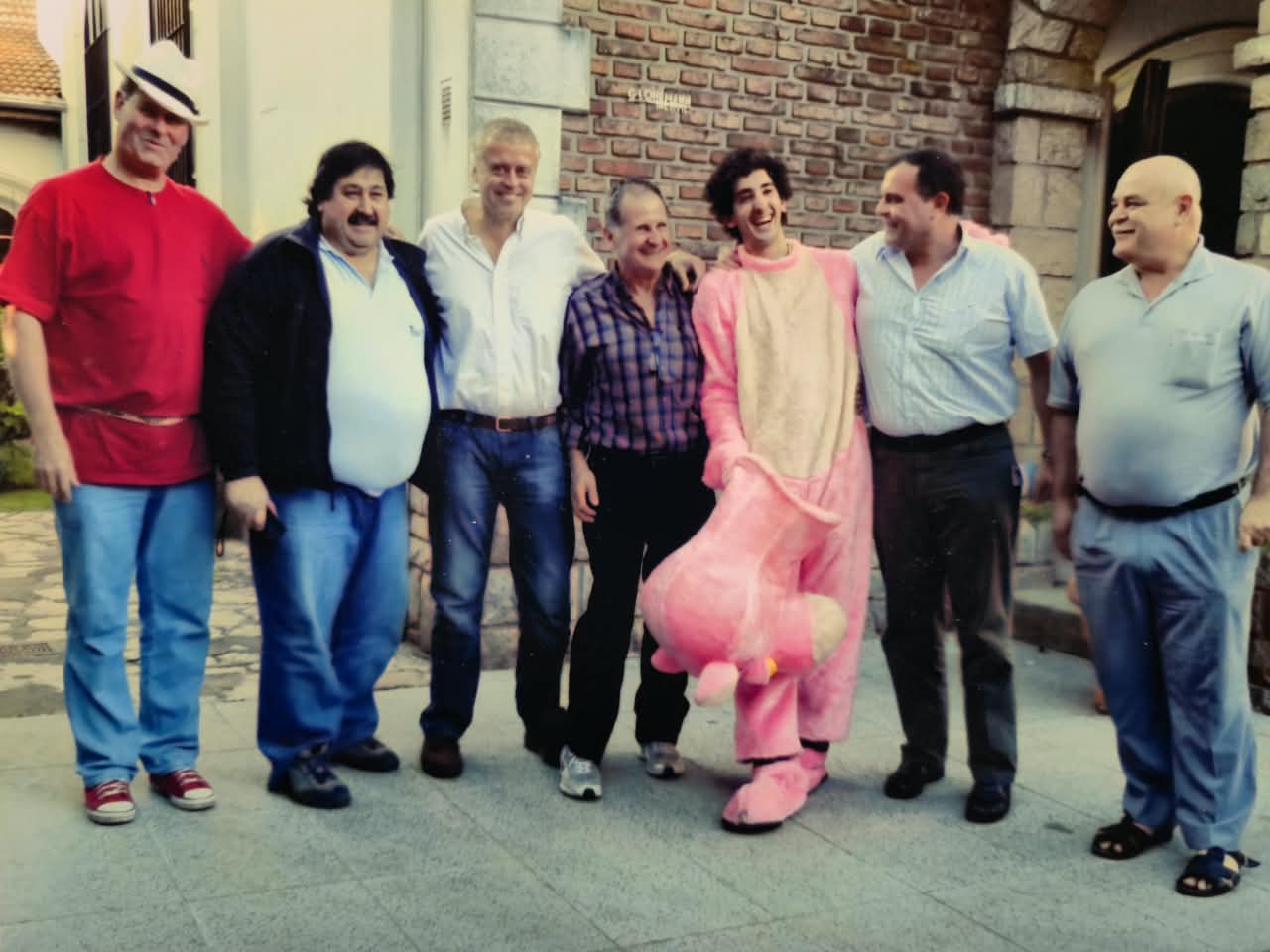 Siete hombres sonrientes de pie en un patio frente a un edificio de ladrillo; uno, en el centro, disfrazado con un traje de peluche rosa