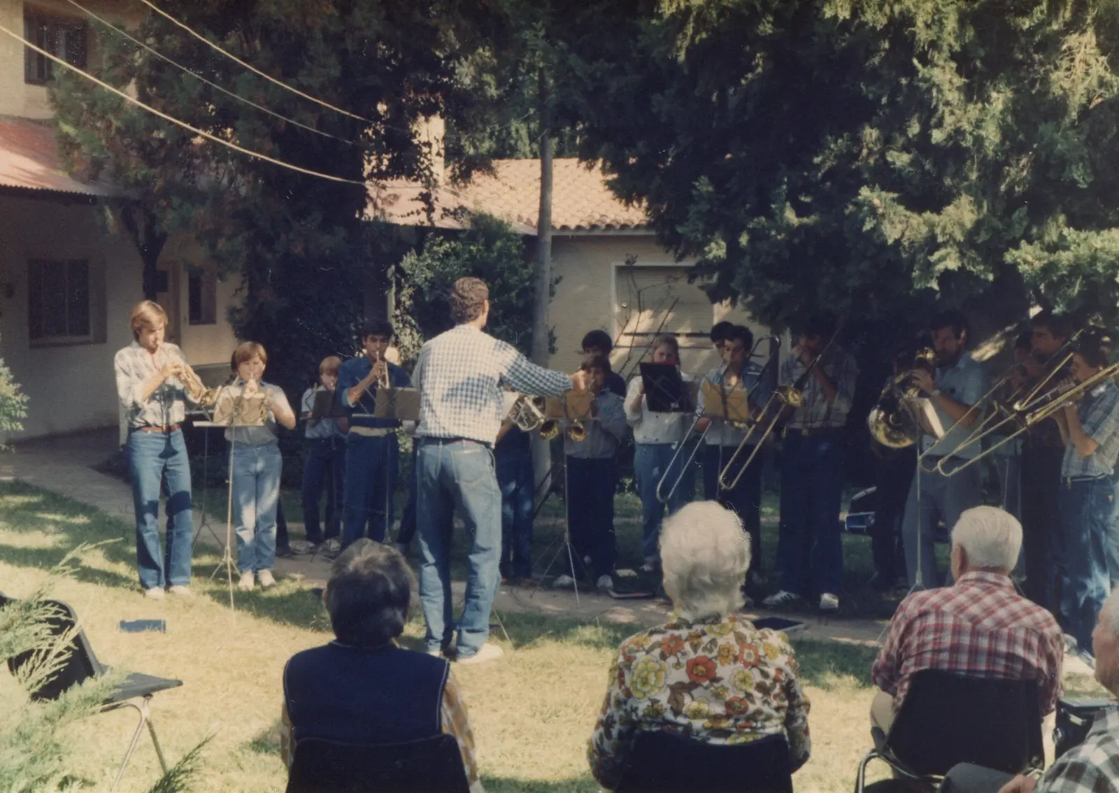 Concierto al aire libre en el jardín de una casa de techo de tejas: unos quince músicos de jean y camisa a cuadros de pie en semicírculo con atriles, y en primer plano tres personas mayores sentadas de espaldas en sillas plegables sobre el pasto