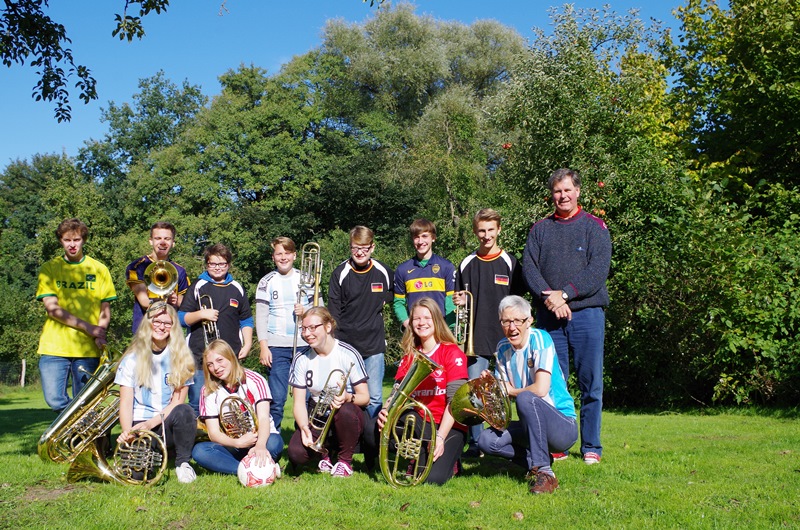 Catorce integrantes del Junger Bläserkreis M-V posando en un prado con camisetas de fútbol y los instrumentos adelante