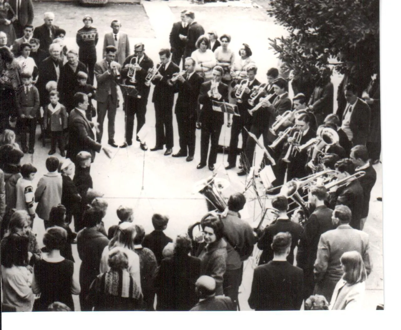 Fotografía en blanco y negro tomada desde arriba: unos dieciocho músicos tocando en semicírculo en un patio, rodeados por completo de gente de todas las edades, con el director a la izquierda dirigiendo con los brazos abiertos