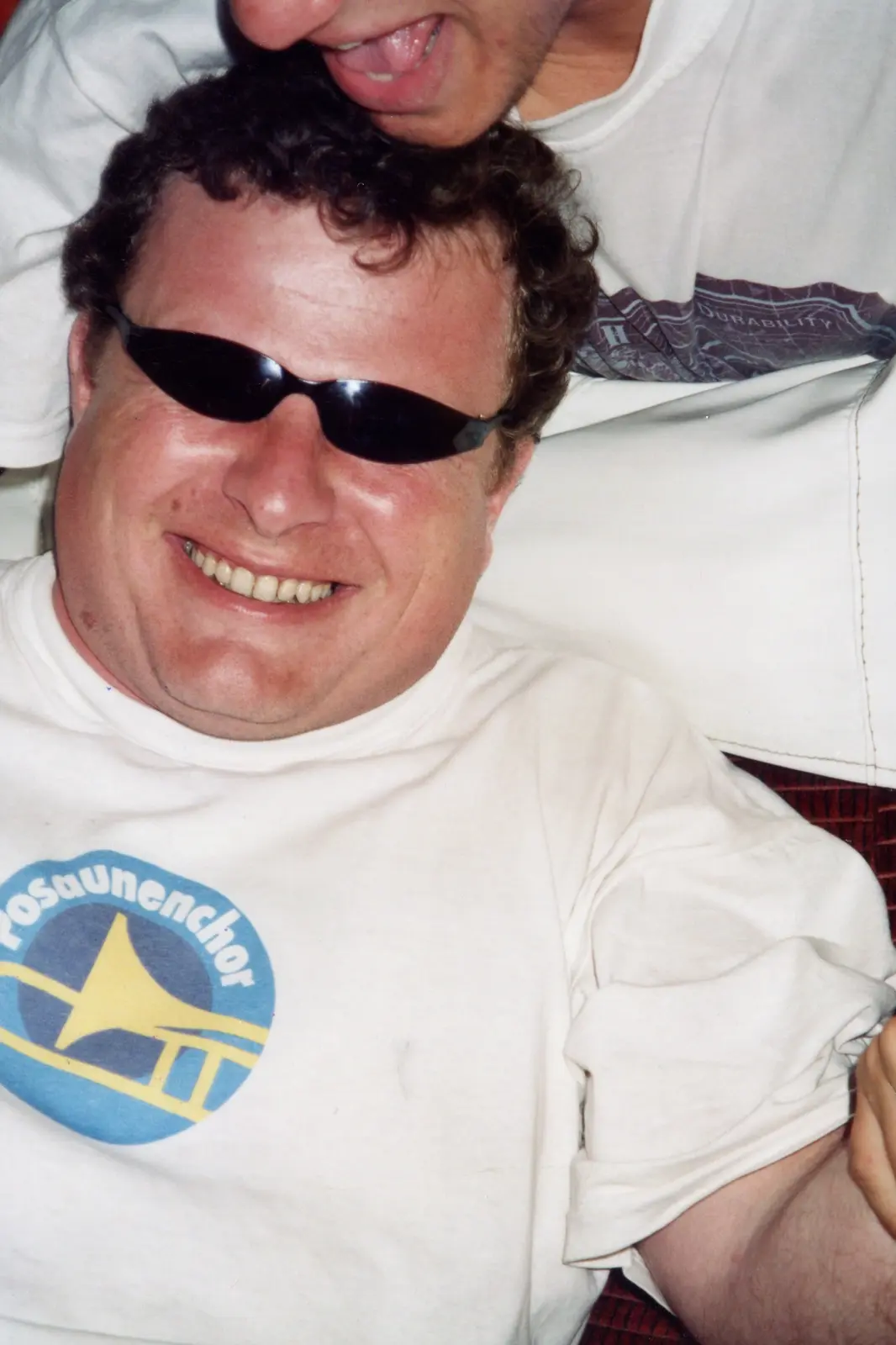 Primer plano sonriente de un hombre con anteojos de sol y una remera del Posaunenchor, con alguien haciendo una morisqueta detrás