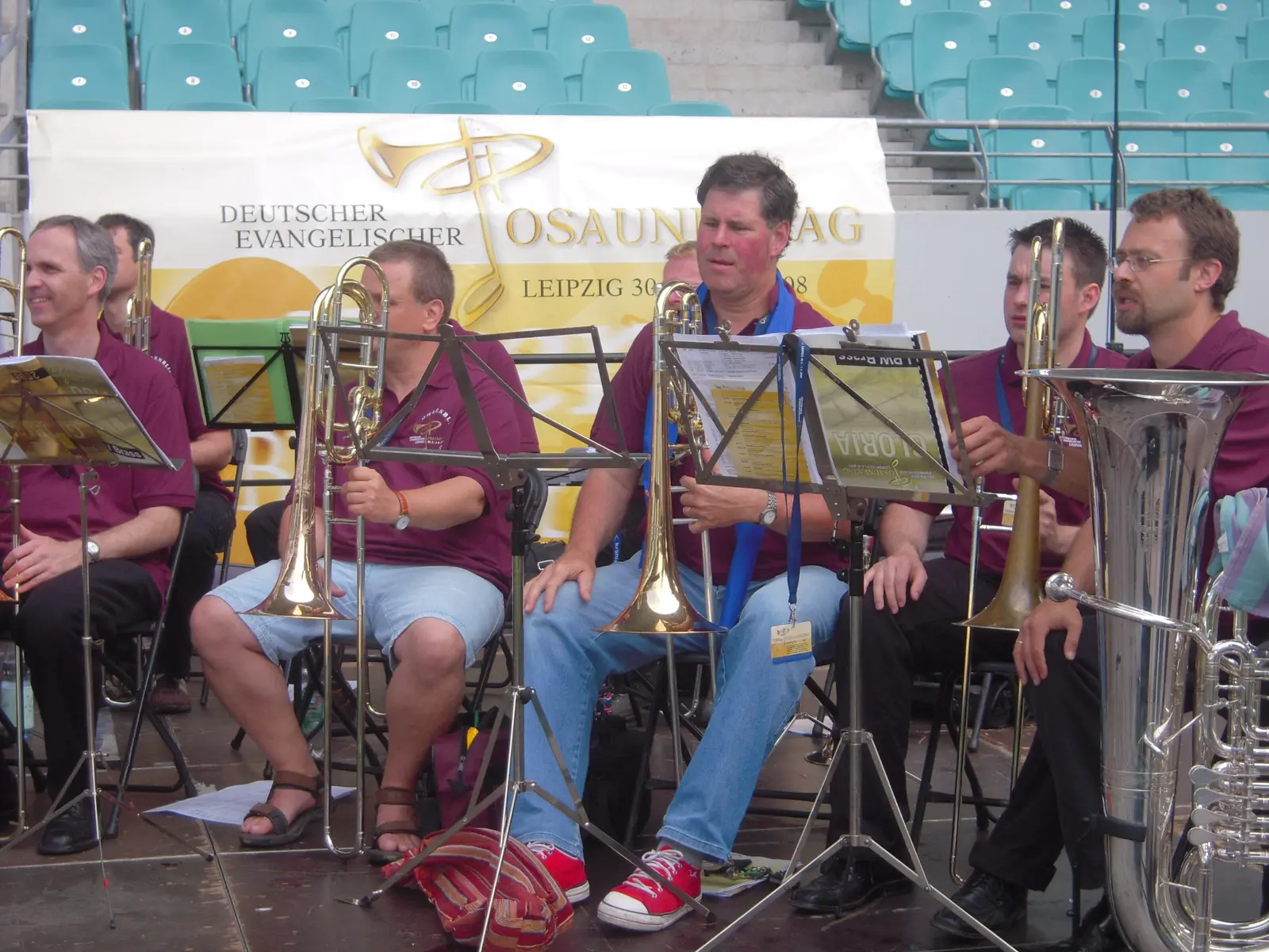 Músicos sentados con trombones y una tuba delante de una lona blanca colgada de la tribuna que dice Deutscher Evangelischer Posaunentag Leipzig 2008, con un logo dorado de trompeta y cruz