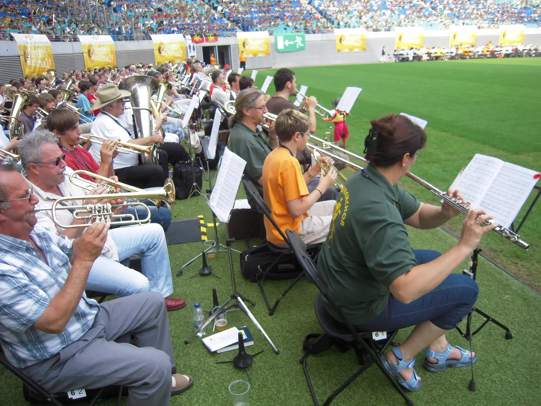 Vista desde la tribuna del estadio de Leipzig con miles de músicos sentados en sillas numeradas sobre el césped