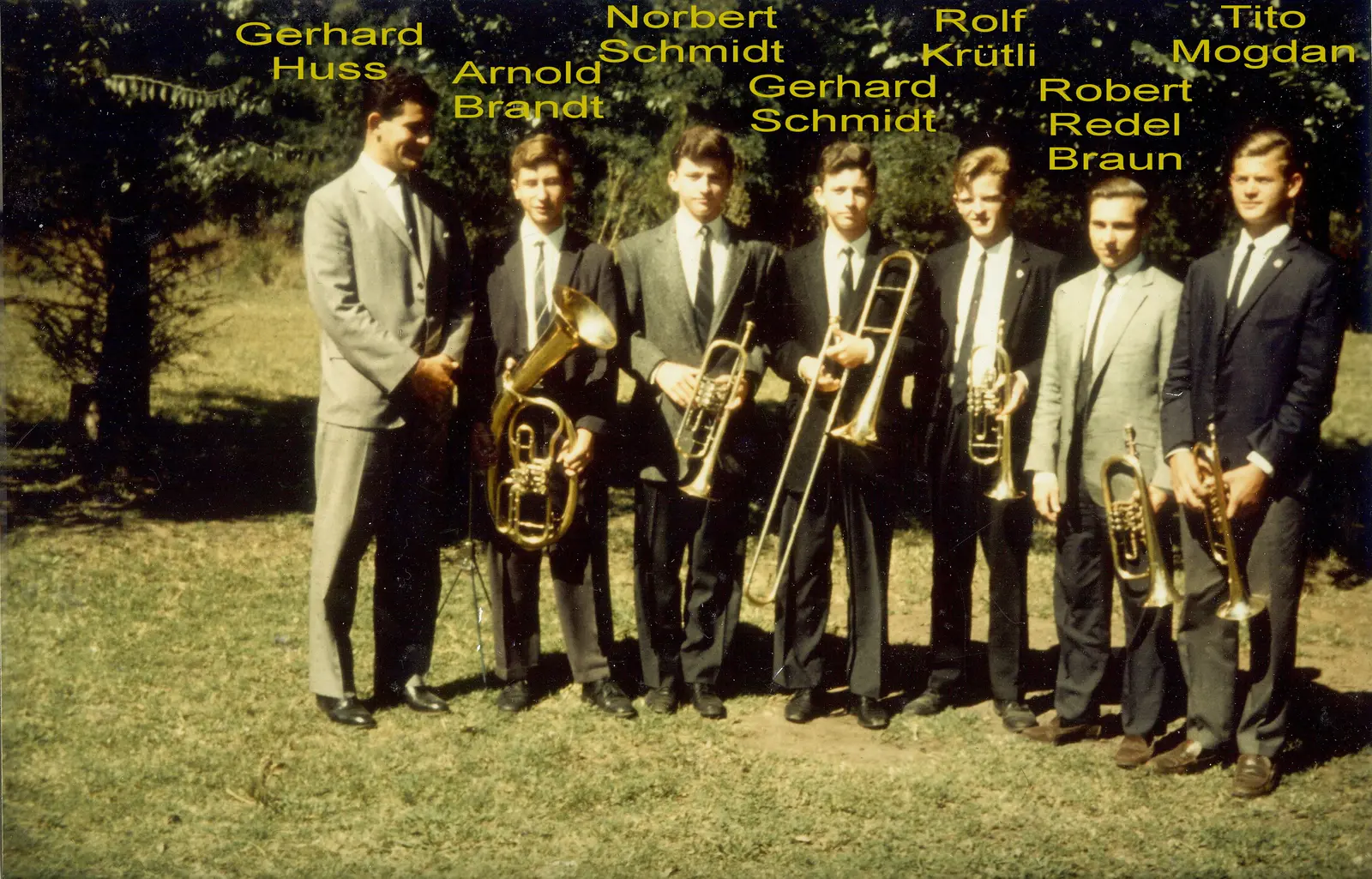 Siete músicos de traje con sus instrumentos en un jardín, fotografía en color de mediados de los años sesenta con nombres rotulados