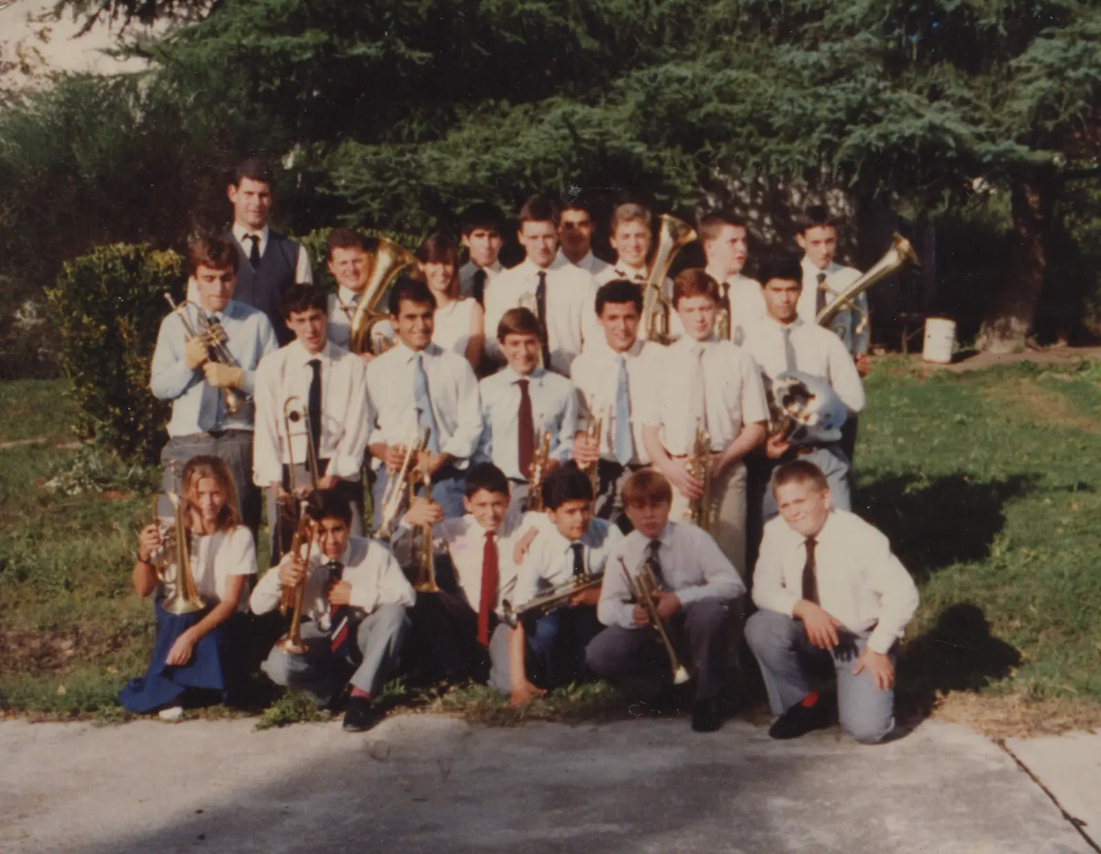 Retrato grupal de unos veintidós músicos sobre el pasto delante de una conífera grande, en tres niveles: parados atrás, parados adelante y arrodillados en el suelo, todos con camisa clara, corbata y su instrumento