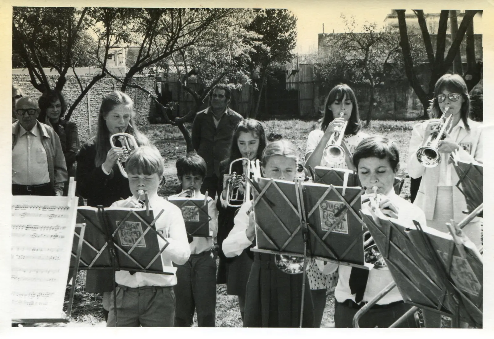 Fotografía en blanco y negro: nueve chicos y chicas tocando trompetas y fliscornos al aire libre en un terreno con árboles pelados y un muro al fondo, frente a atriles con carpetas de tapa impresa