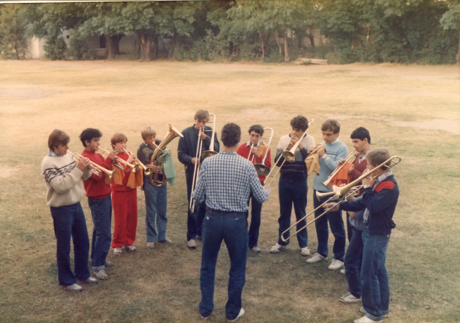 Once adolescentes en semicírculo sobre el césped tocando trompetas, fliscornos, un bombardino y trombones, sin ningún atril a la vista, con el director de espaldas en el centro con camisa a cuadros
