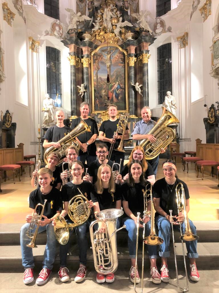 Un grupo de músicos con trompetas, cornos, trombones, bombardinos y tubas, de pie y sentados en los escalones del presbiterio de una iglesia barroca, delante de un retablo con columnas oscuras, ángeles blancos y un cuadro de la crucifixión