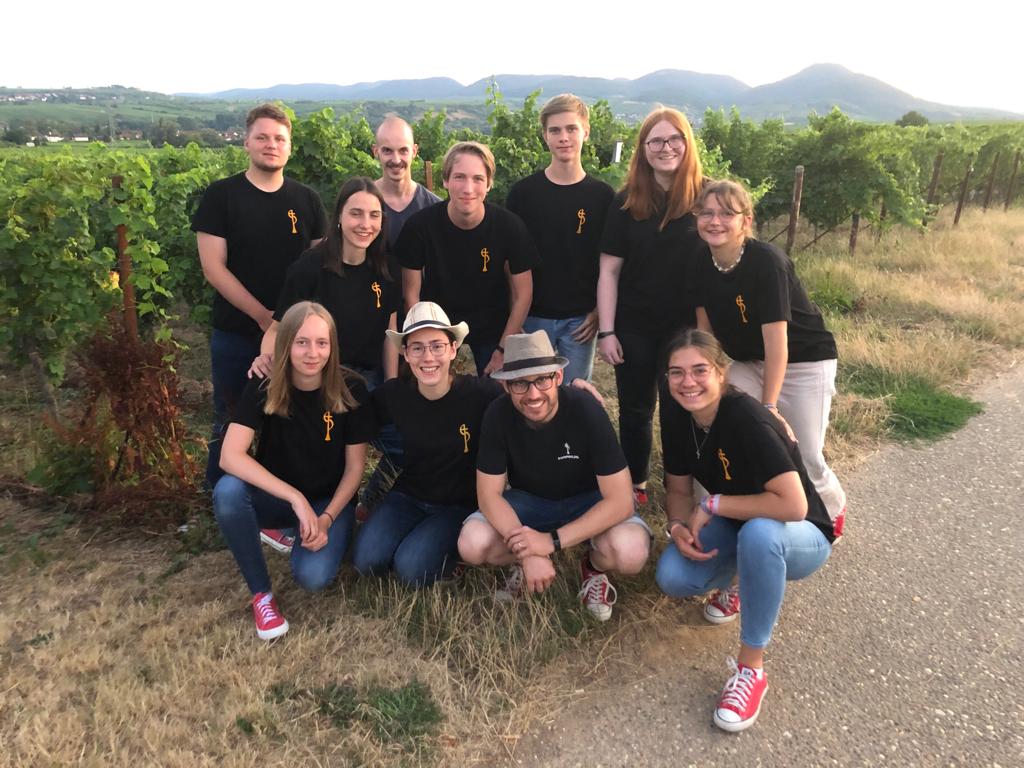 Un grupo de jóvenes y adultos con remeras negras, de pie y en cuclillas al costado de un camino, entre las hileras de un viñedo; al fondo, viñas hasta el horizonte y una cadena de cerros bajos