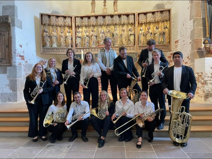Un conjunto de bronces con trompetas, trombones, cornos, bombardinos y una tuba, de pie y sentados en los escalones del presbiterio de una iglesia, delante de un retablo dorado tallado