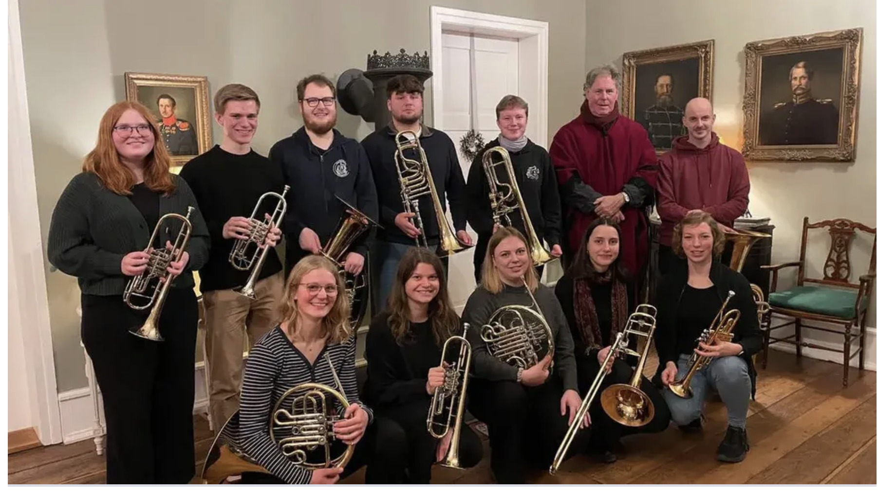 Doce músicos de pie con sus instrumentos de bronce —trompetas, trombones de vara, cornos y una tuba— en una sala de paredes claras con grandes retratos al óleo enmarcados colgados detrás de ellos