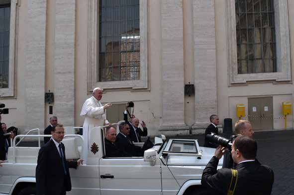 El papa Francisco de pie en el papamóvil blanco recorriendo la plaza, con un fotógrafo delante