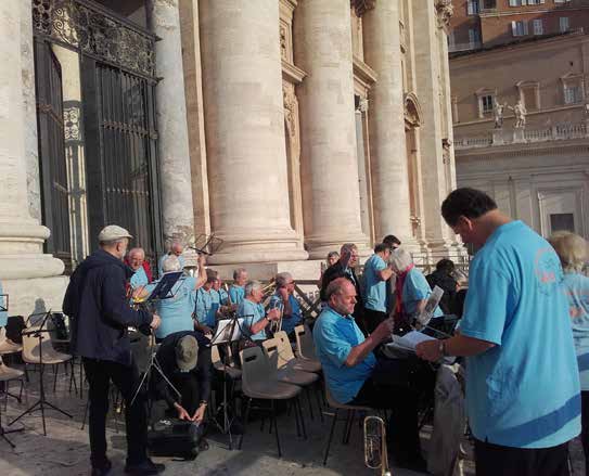 Los músicos de camisa celeste tocando en su puesto de la Plaza de San Pedro, con atriles y tubas