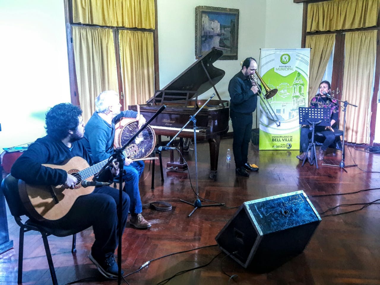 Cuatro músicos en un salón con piano de cola: un guitarrista y un percusionista con bombo sentados, Pablo Fenoglio tocando el trombón de pie y una cantante sentada ante un micrófono