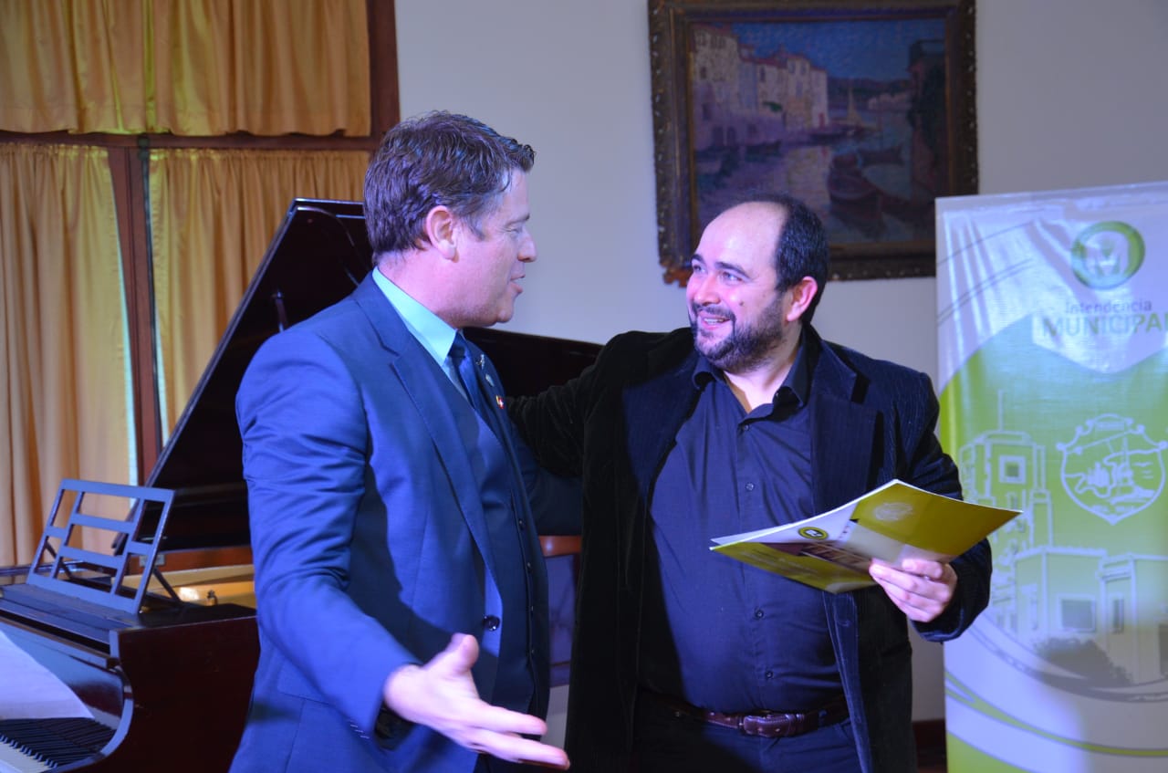 Pablo Fenoglio, a la derecha, de camisa y saco negros, sostiene una carpeta amarilla y conversa sonriente con un hombre de traje azul junto a un piano de cola
