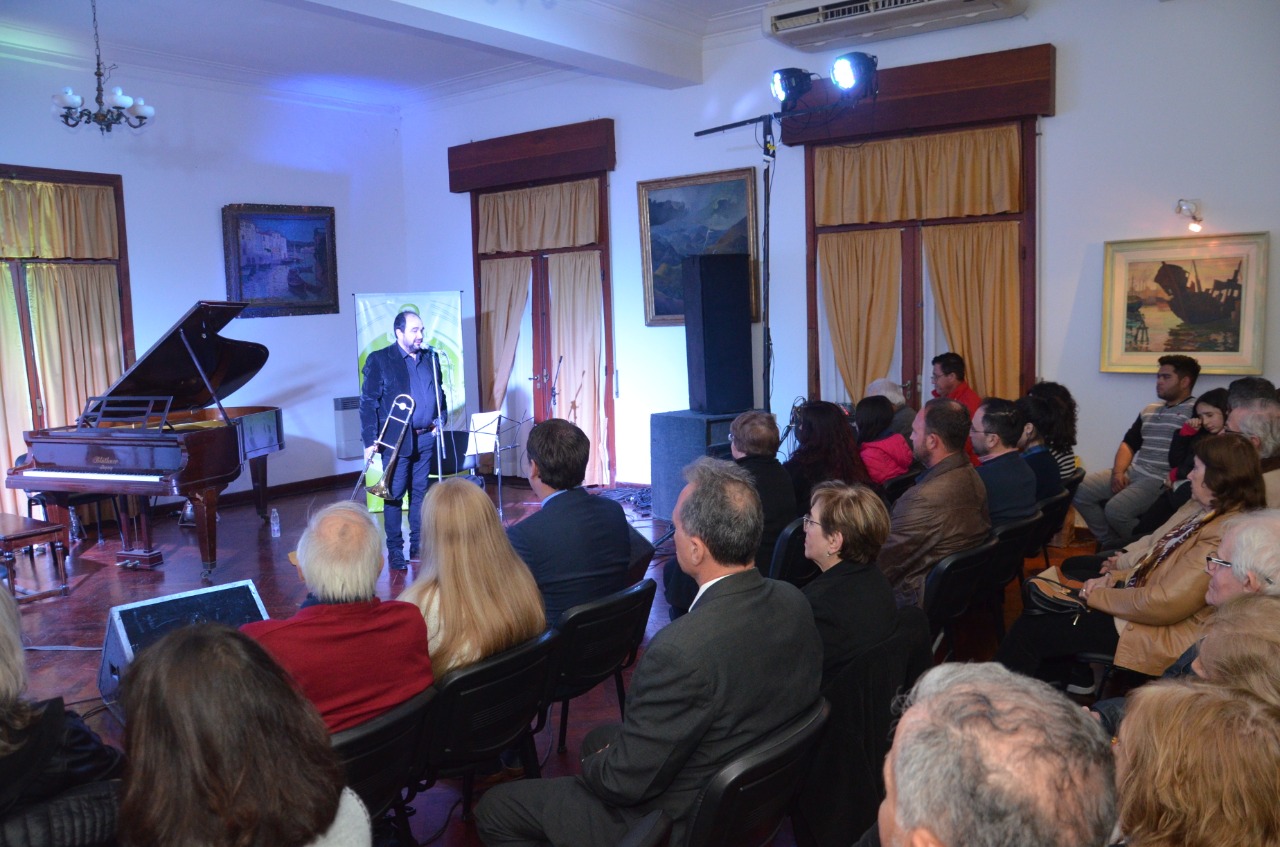 Sala de actos con público sentado de espaldas; al frente, junto a un piano de cola y un banner con su retrato, Pablo Fenoglio habla al micrófono con el trombón en la mano