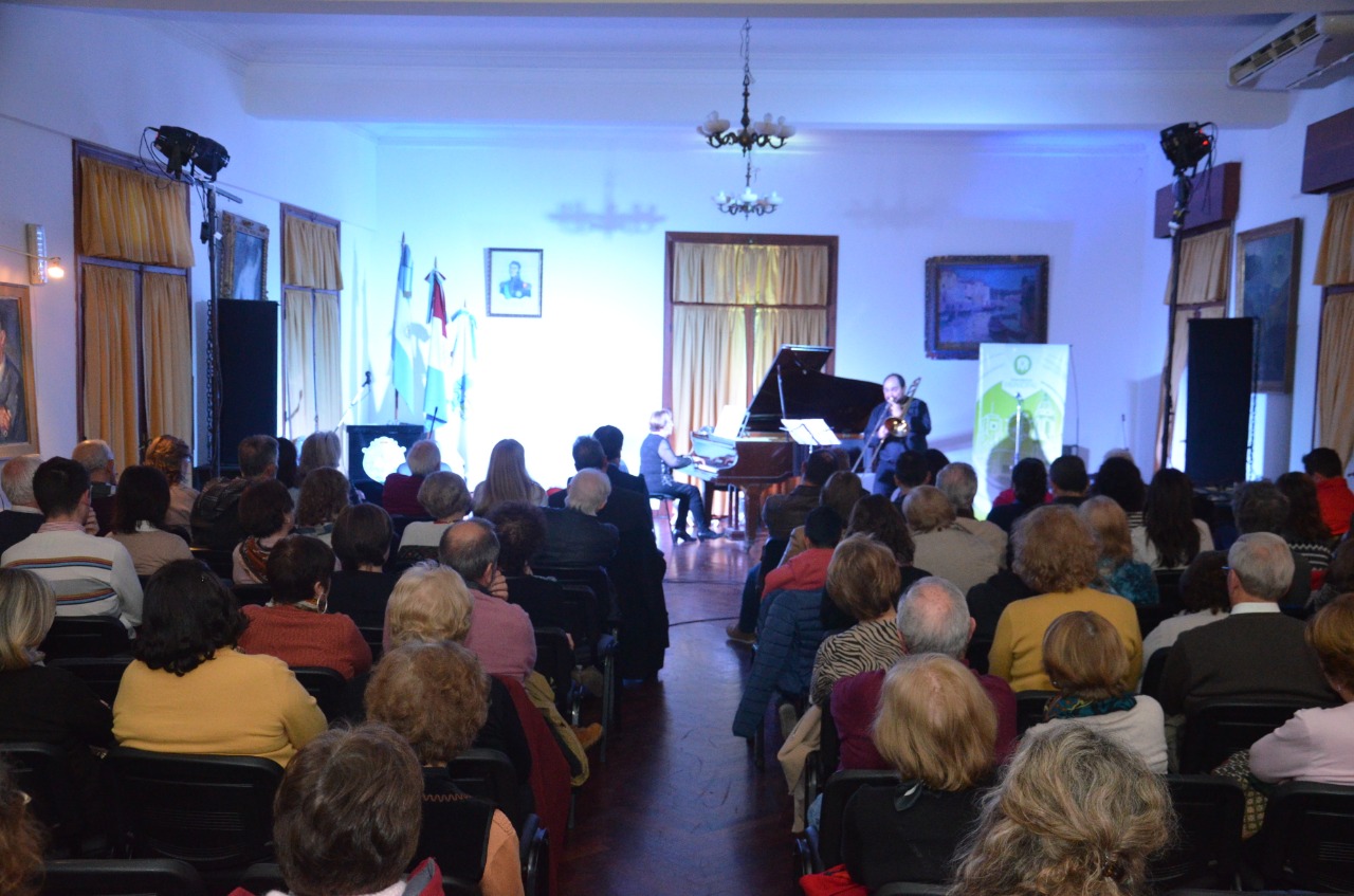 Vista general del salón con el público de espaldas llenando las sillas; al fondo, iluminados, una pianista al piano de cola y Pablo Fenoglio de pie con el trombón