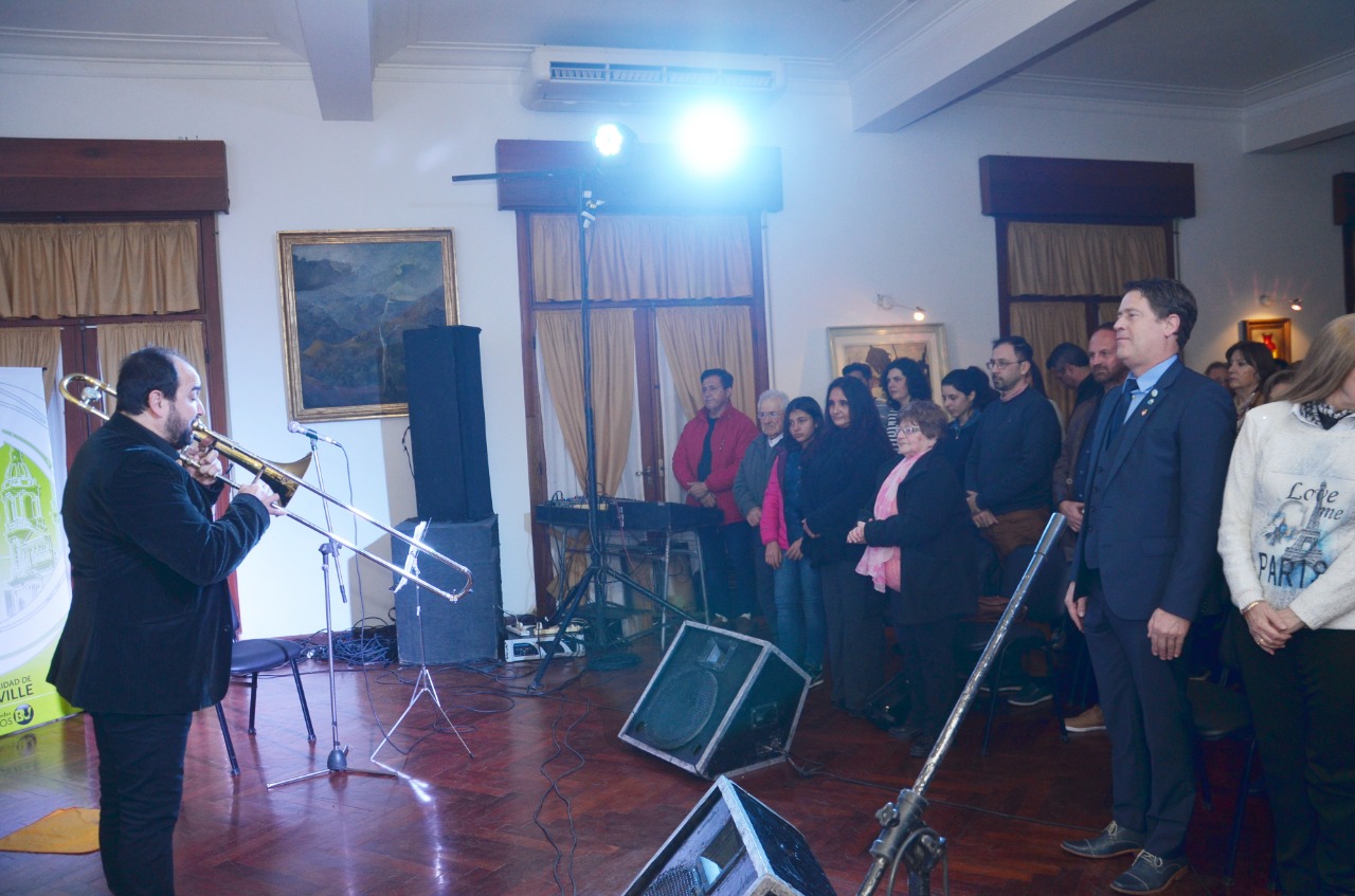 Pablo Fenoglio, de perfil y de negro, toca el trombón frente a un micrófono mientras el público permanece de pie mirándolo