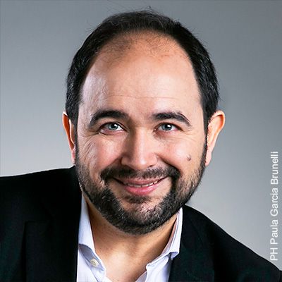 Retrato de Pablo Fenoglio sonriendo, de saco oscuro y camisa blanca, con el pelo corto y barba, sobre fondo gris