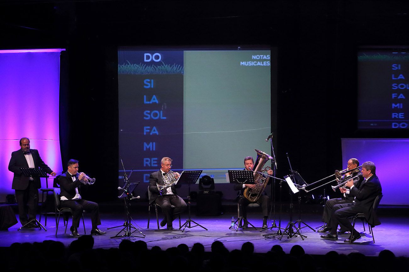 Totale des Quinteto Bronces del Plata auf der Bühne im blauen Licht, mit einer Leinwand, die die Namen der Notenstufen projiziert, und dem Tubisten spielend in der Mitte