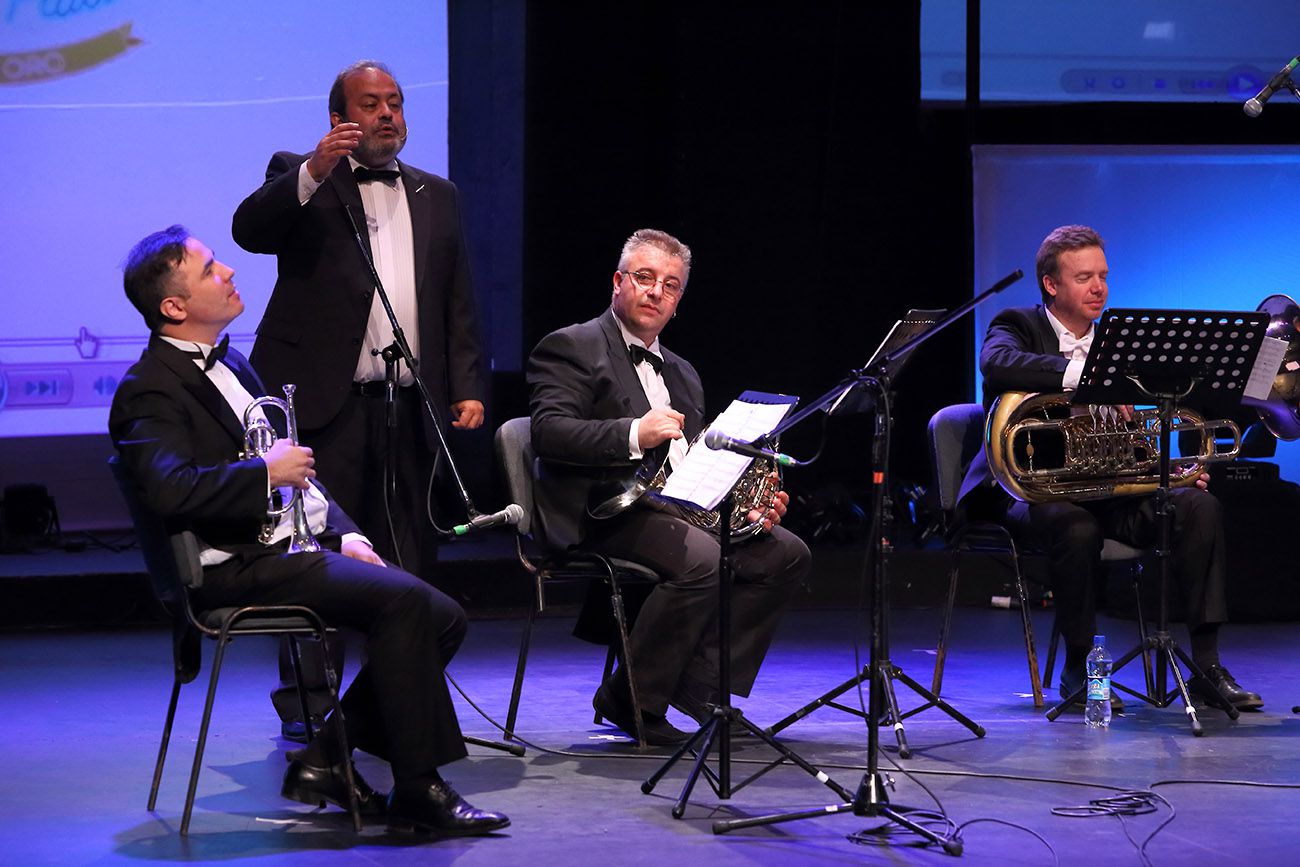 Pedro Pulzován sitzt rechts auf der Bühne, die Tuba auf den Knien, das Gesicht frei sichtbar, im blauen Licht, daneben ein Trompeter und ein Hornist sowie ein stehender Schauspieler