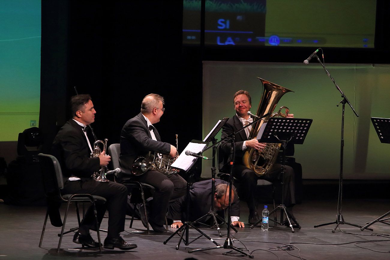 Pedro Pulzován sitzt rechts auf der Bühne mit einer goldenen Tuba auf den Knien und lächelt zu seinen Kollegen im Frack hinüber, während ein Schauspieler am Boden liegend einen Gag spielt