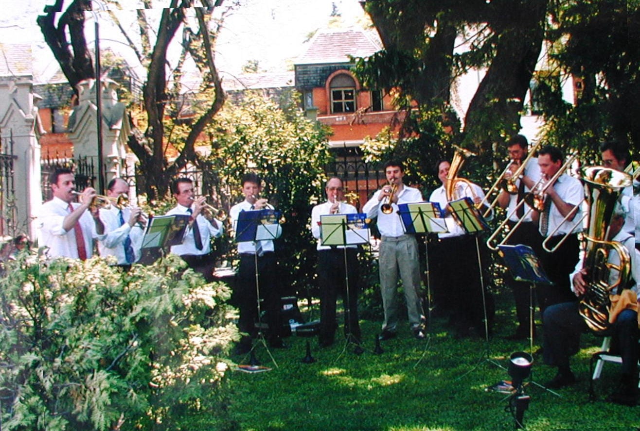 Once músicos de traje —camisa blanca y corbata— tocan de pie al aire libre sobre el césped, en fila abierta: cinco trompetas, dos trombones de vara, un corno y una tuba, con atriles negros y carpetas de partituras azules y verdes; detrás, una casa de dos plantas de ladrillo a la vista con techo de tejas y una ventana en arco, y a la izquierda una reja negra con pilares de mampostería